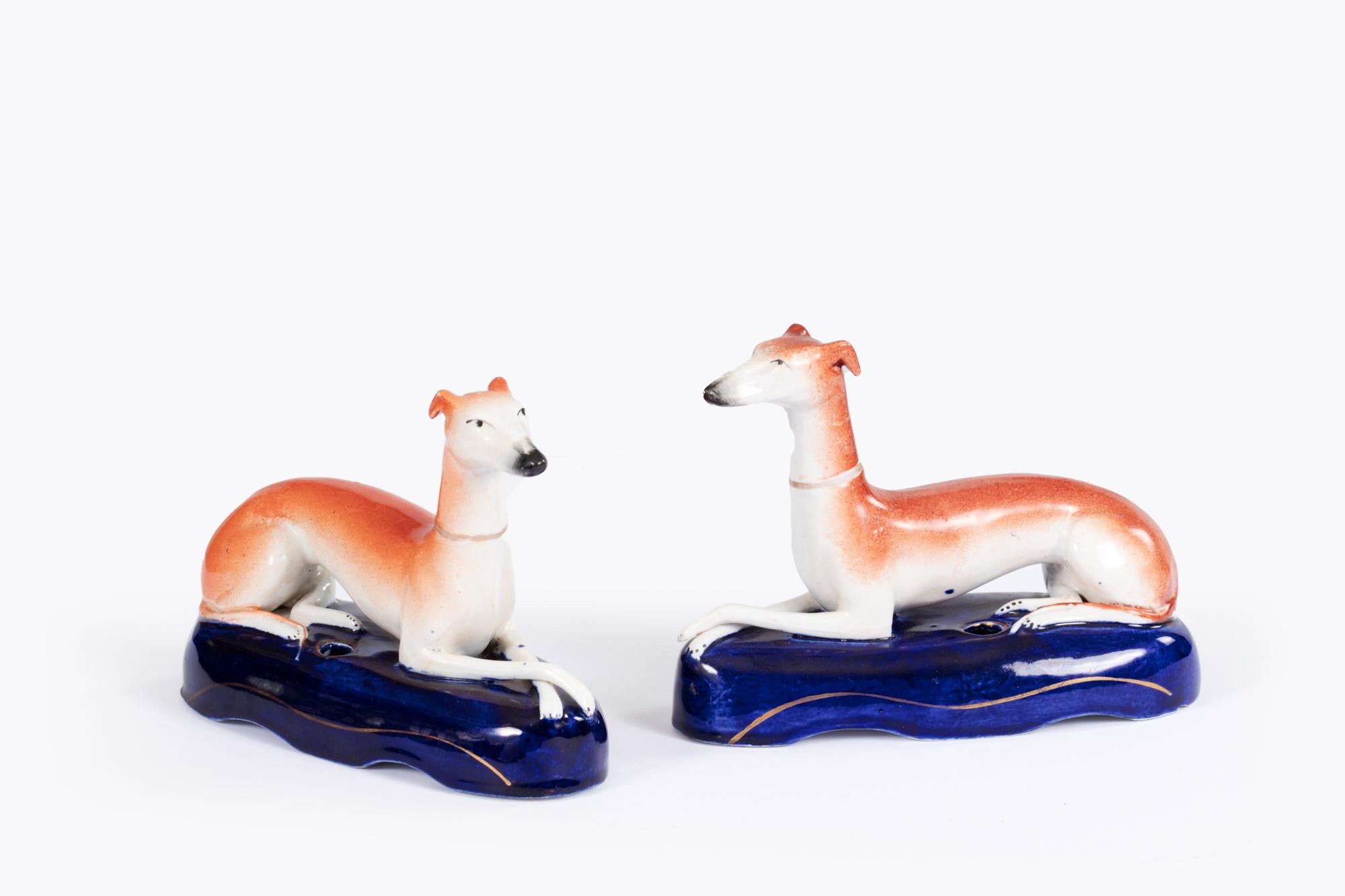 19th Century pair Staffordshire pottery pen stands in the form of resting greyhounds. The pair are poised with crossed paws laying on a cobalt blue scalloped pedestal with gold gilt trim. Circa 1860.