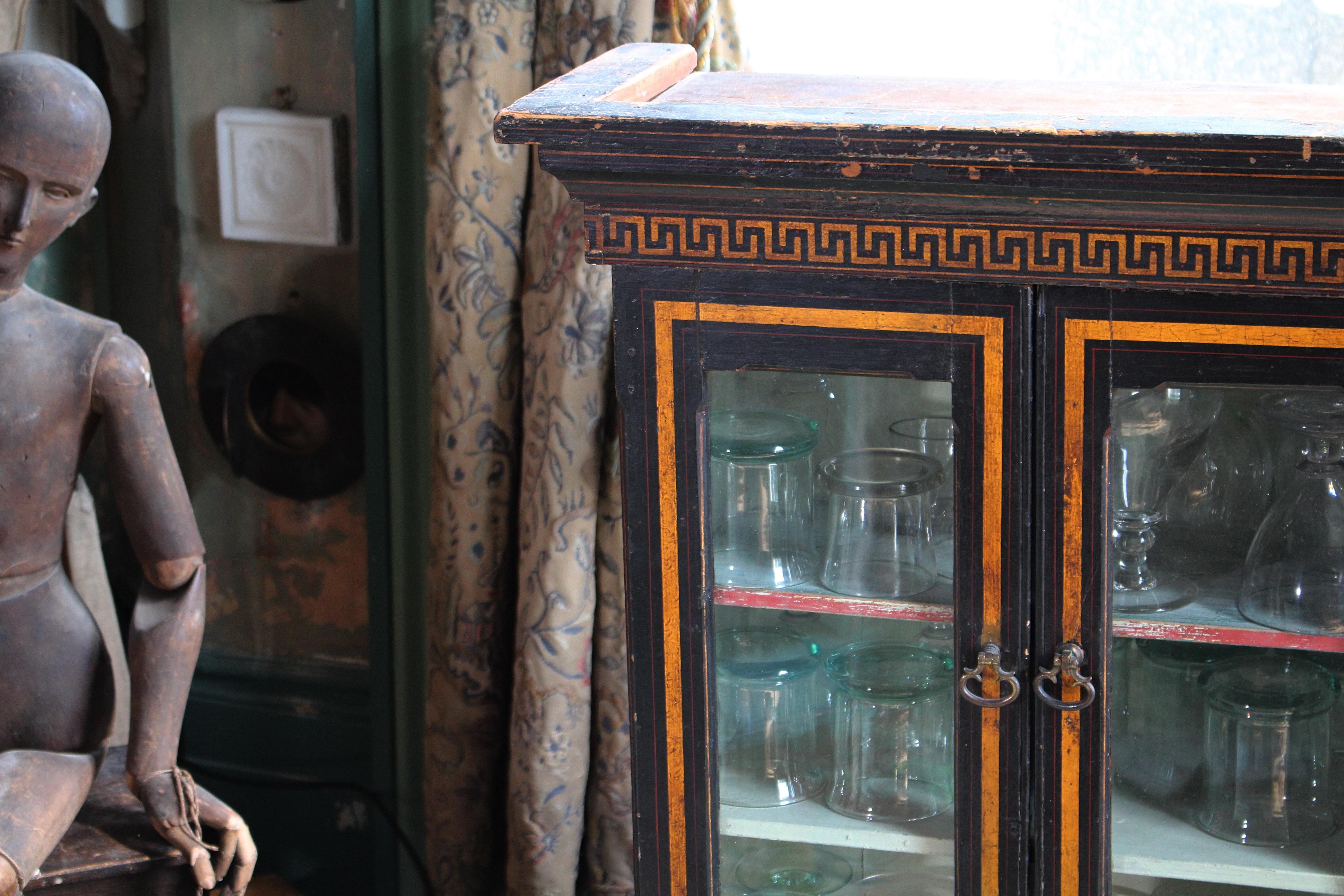 A mid to late 19th century ebonised glazed wall hanging display cabinet, with two glazed doors and two shelves within.

The cabinet decorated with a gilt and ruby red lineal design, along with a bold greek border at the bottom of the pelmet.

The
