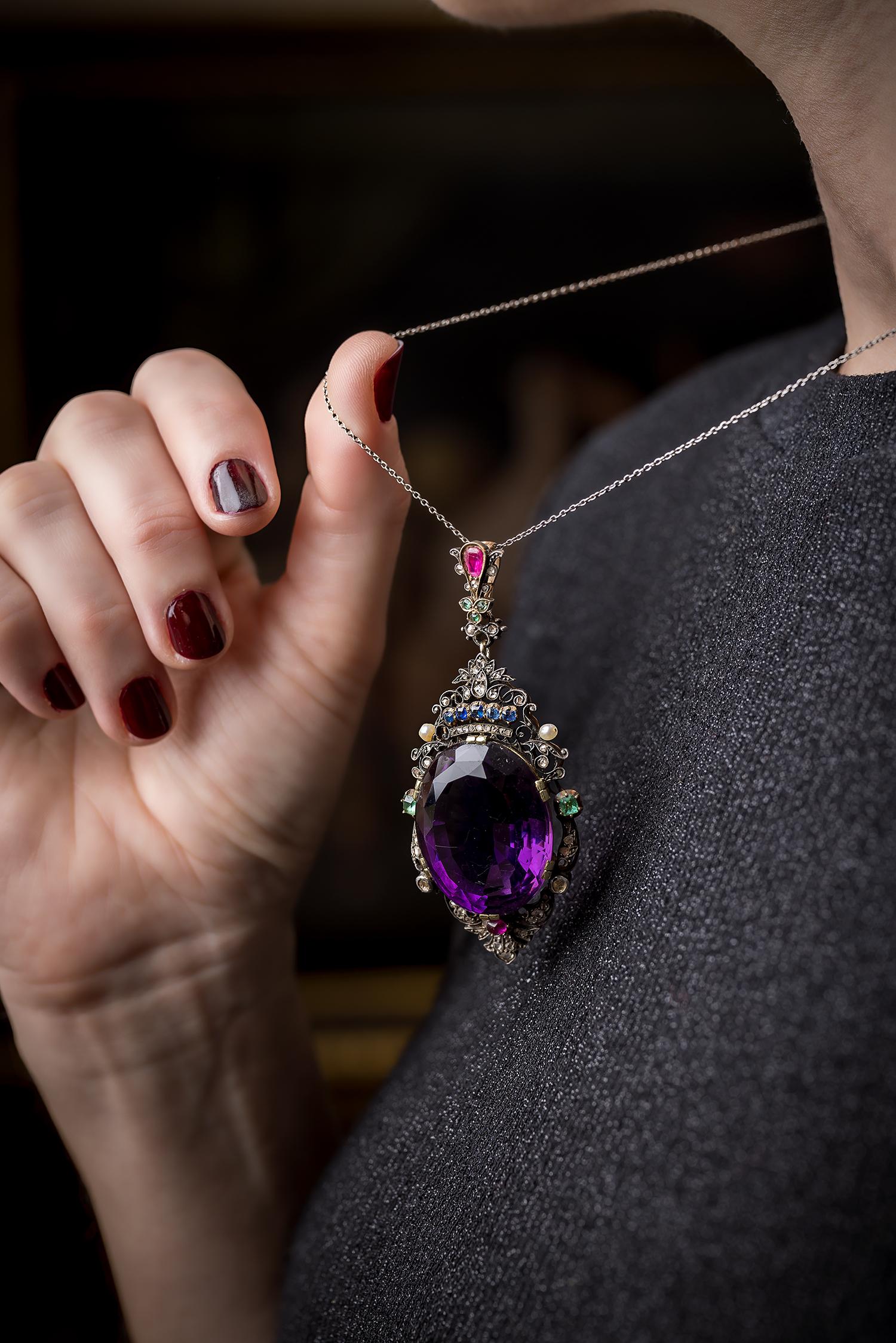 An impressive 45 carat oval faceted and deeply saturated amethyst gracefully set framed in a scrolling foliate under a sapphire crown and embellished by rubies, emeralds, sapphires, diamonds and oriental pearls, Masterfully hand fabricated in 14
