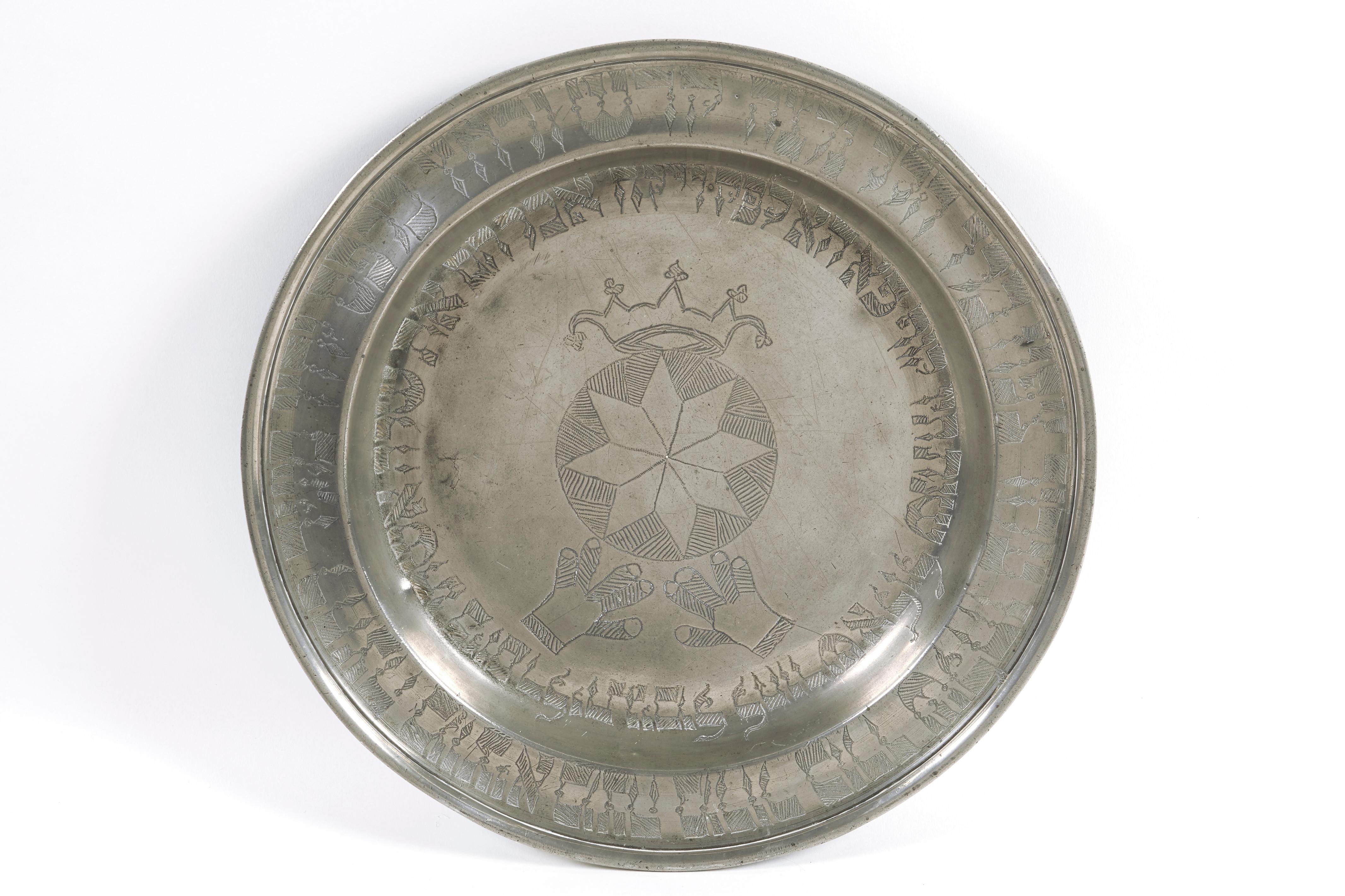 Assiette en étain allemande pour la Pâque, datée de 1825.
La plaque à bord relevé est gravée en hébreu de l'ordre du Seder de Pessah. Au centre, un motif géométrique, entouré de texte hébreu pour la date et le nom du propriétaire, surmonté d'une