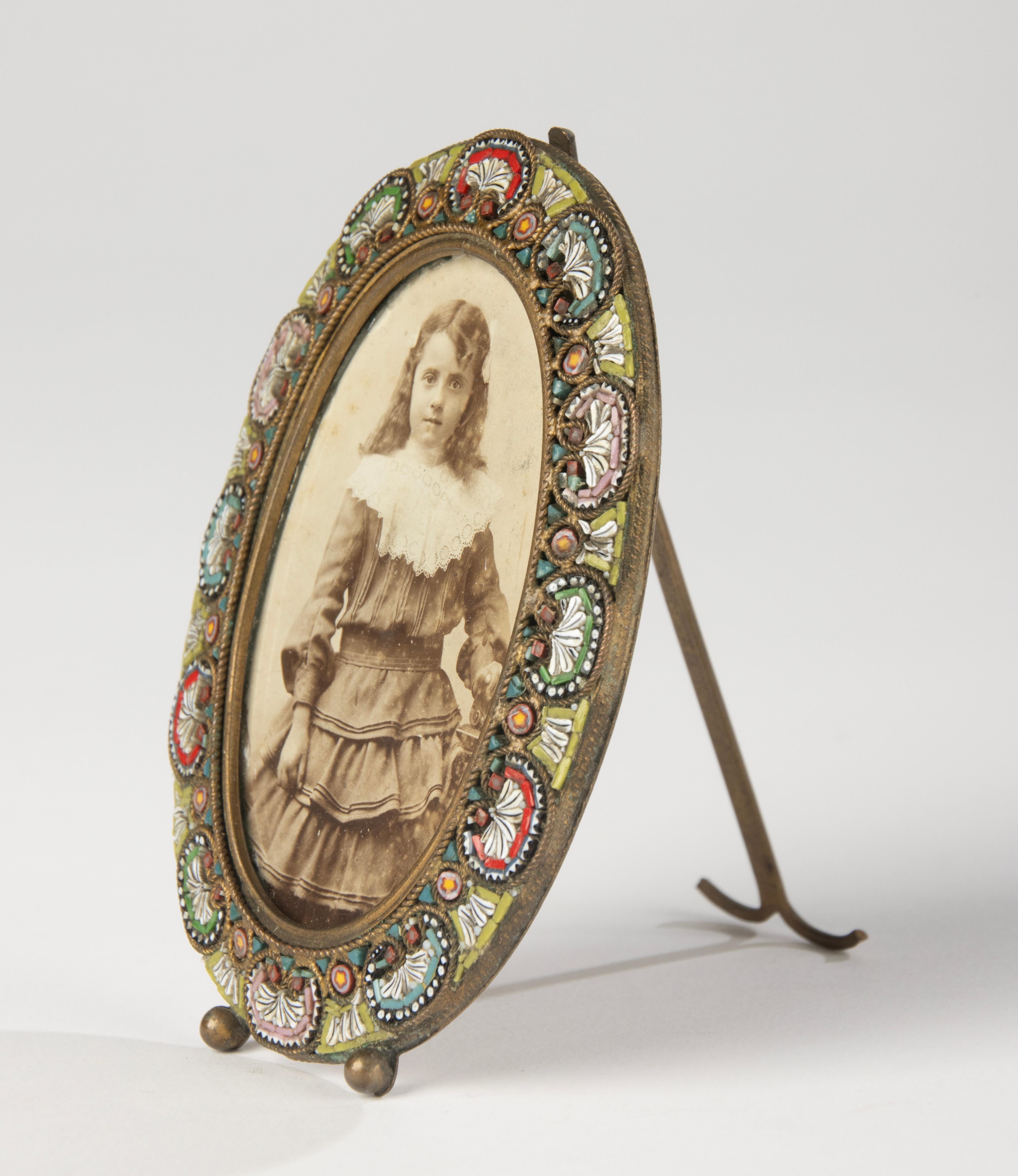 Charming small antique photo frame, crafted around 1890-1900, made of brass and beautifully inlaid with micro glass mosaic in delicate colors. The oval frame is finely detailed and features intricate floral patterns, showcasing the high level of