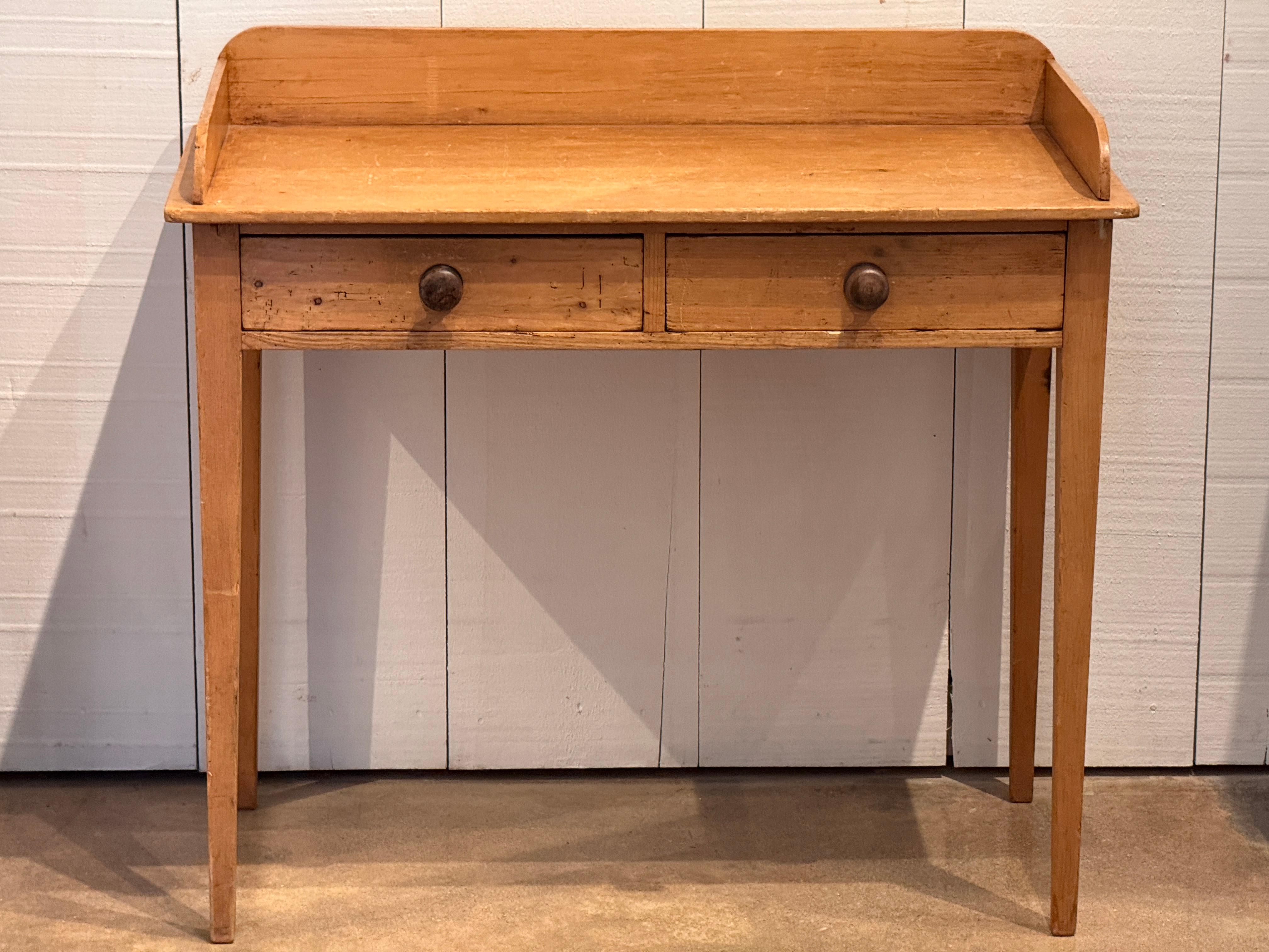 Table en pin avec un plateau à trois côtés en forme de galerie, deux tiroirs et des pieds fuselés.
Les tiroirs sont assemblés à queue d'aronde et dotés de poignées en bois tourné.
Les surfaces présentent une usure et des marques d'outils