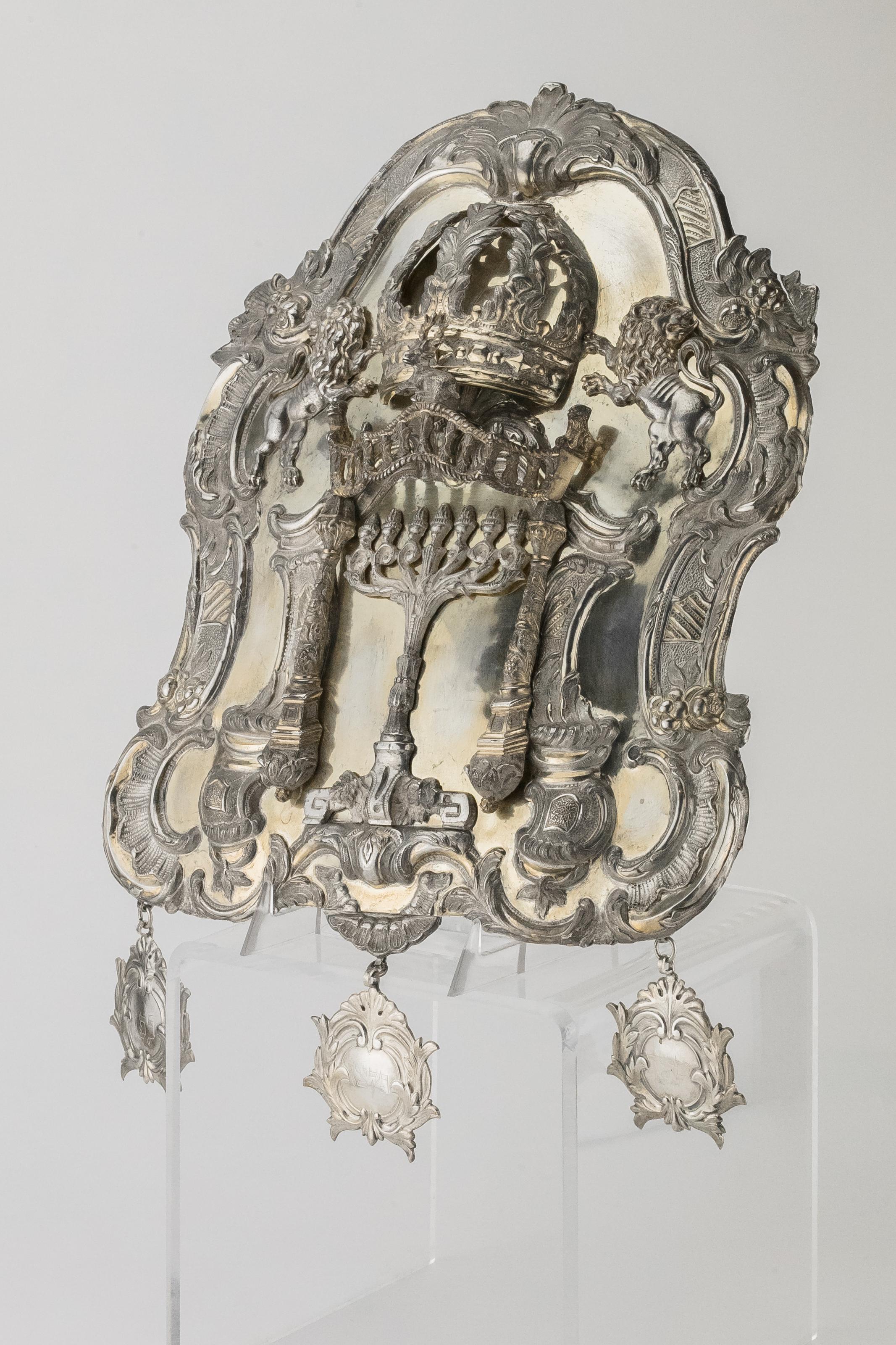 Parcel-gilt silver Torah shield, Warsaw, Poland, 1836.
Of a cartouche form applied with a Menorah under a columned canopy with a crown and lions above, the canopy applied with a stag and two lions, with chased rococo borders, hung with three