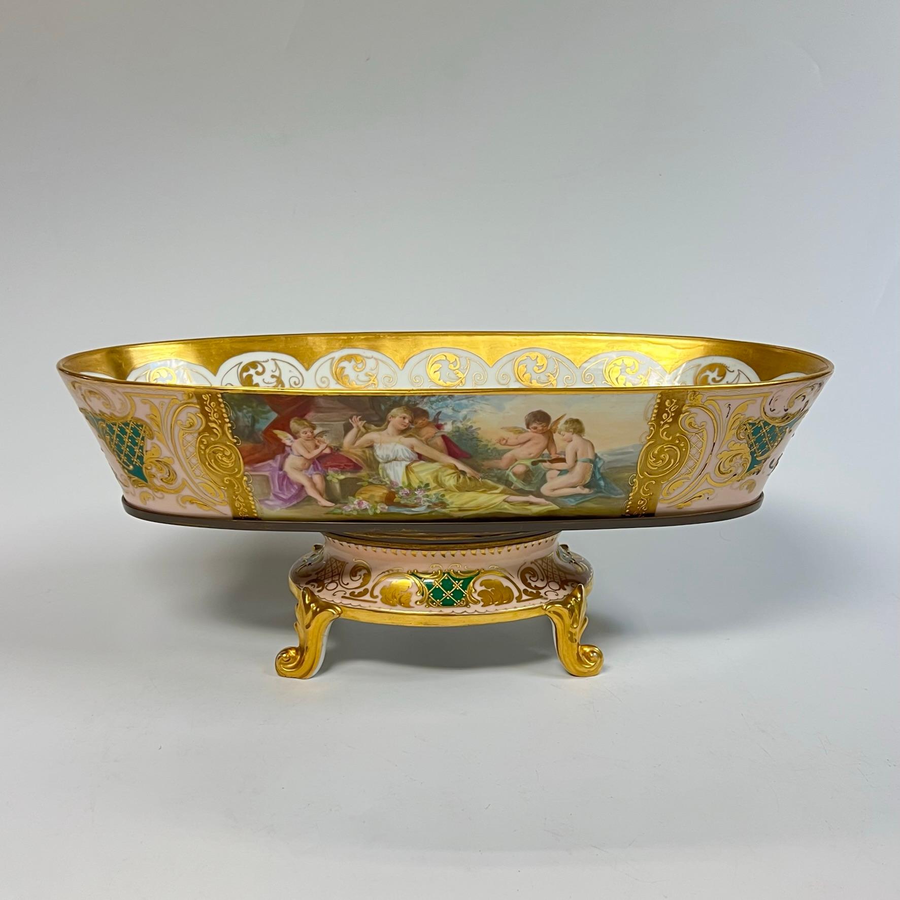 Oval shaped jardiniere or centerpiece bowl on footed stand from Royal Vienna.  6 1/2 by 16 3/4 by 8 1/4 inches, with gilt and hand-painted cartouches of Roman mythological scenes depicting goddesses and Cupid engaged in music and dance.  With Royal