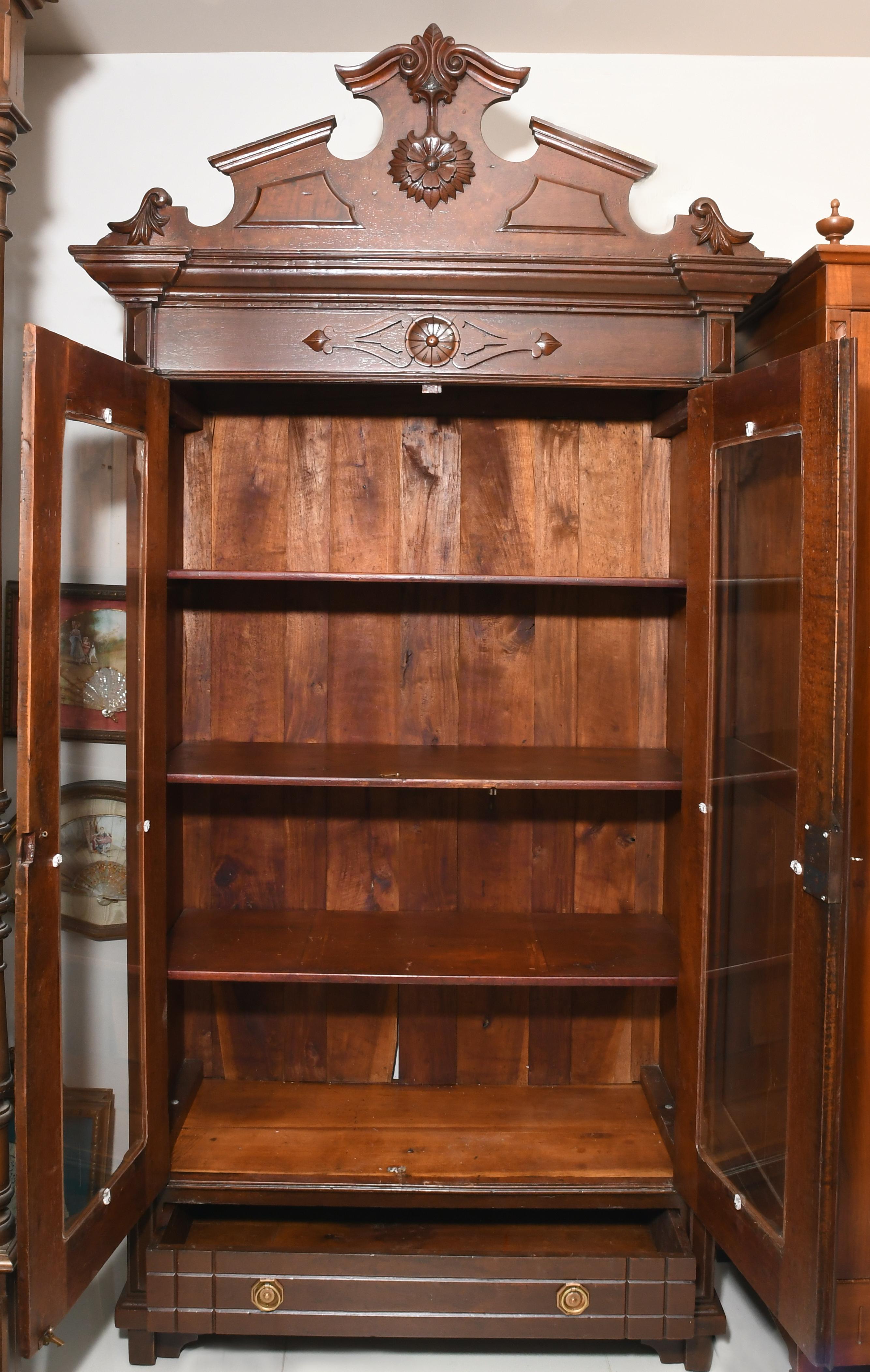 The cabinet has an open arched pediment with a carved crest adorned with a top fleur-de-lis with a pendant flower. A detail of two scrolls holds the fleur-de-lis. There is a carved rosette and two hearts in the center of the cornice. There are two