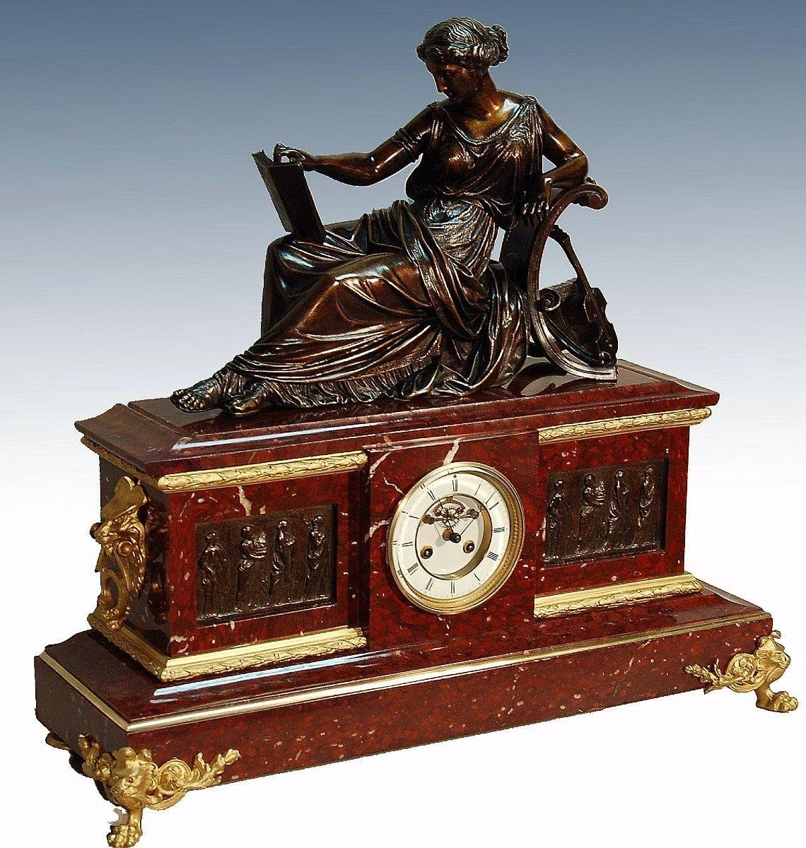 Important clock in royal red marble, surmounted by a patinated bronze sculpture, a woman lying on a couch, she is dressed in antique clothes, she holds a music score in her hand. The theme of this sculpture is music. The red marble is decorated with