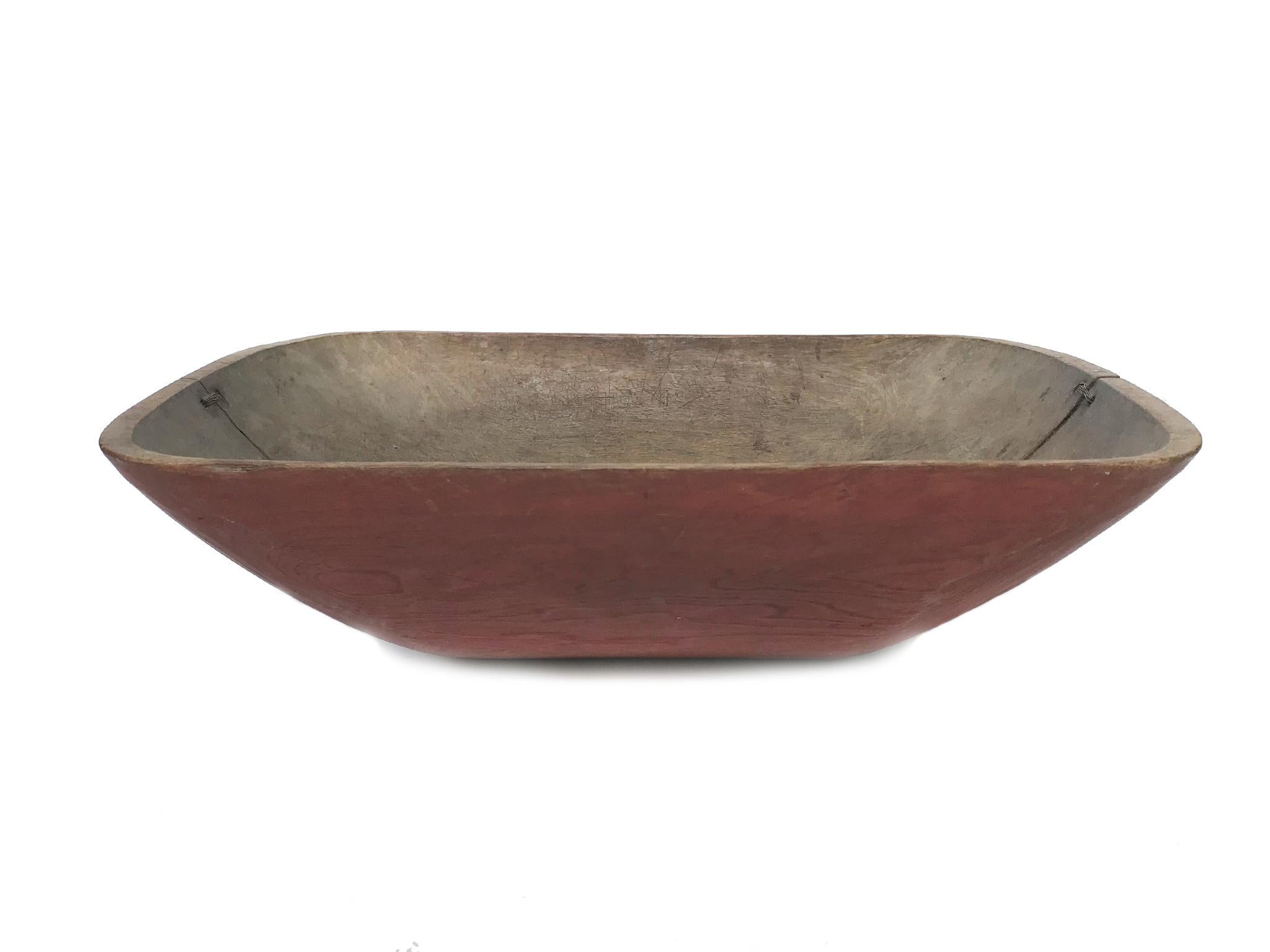 An antique wooden bowl. Hand-carved with a coat of pale red paint on the exterior. The bowl is an artifact that has aged beautifully. Its textures and wear make it a special, historical object. Noteworthy details include the two stitching at either