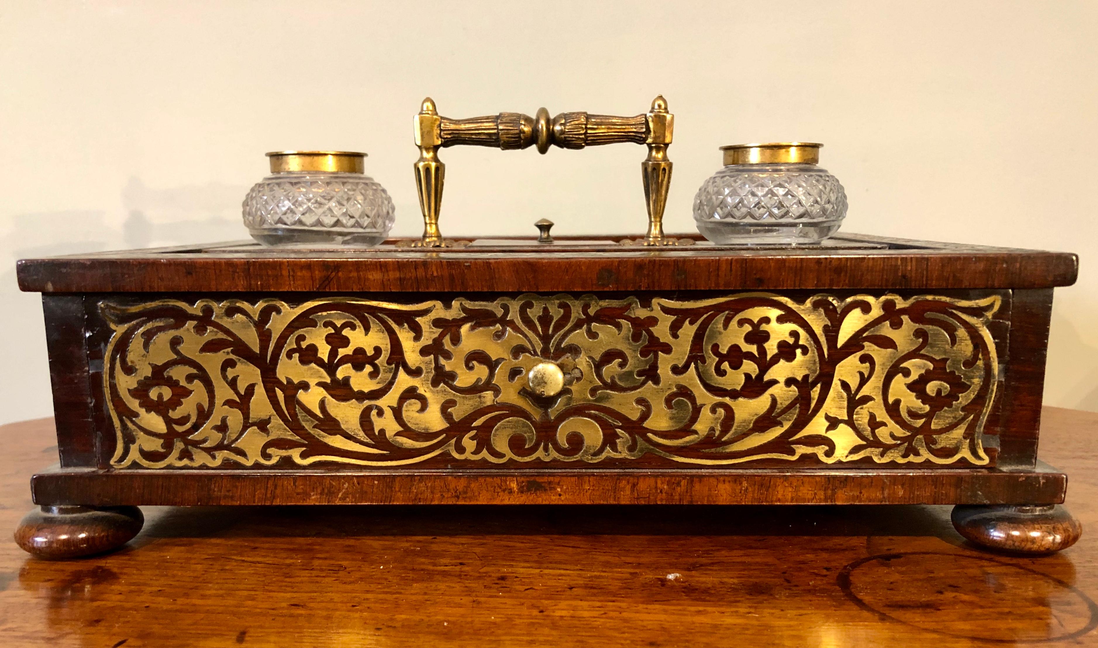 A very handsome Regency period brass inlaid rosewood pen and decoratively cut glass inkwell stand with large central letter drawer and small centre lidded compartment below the ornate brass handle with pen trays to each side. Standing on four small