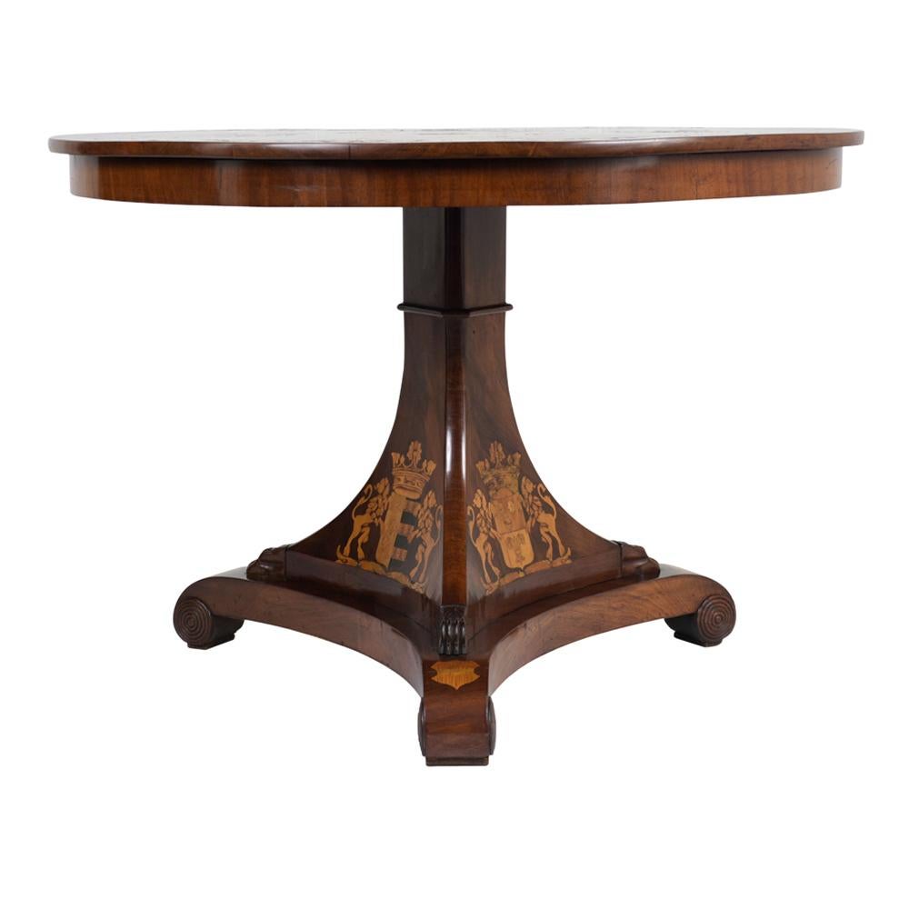 This Circa 1880s English Regency Center Table is made of wood finished in a rich mahogany color with a lacquered finish. The table is ornately adorned with marquetry details of different coat of arms flanked by lions and floral/leafy accents along
