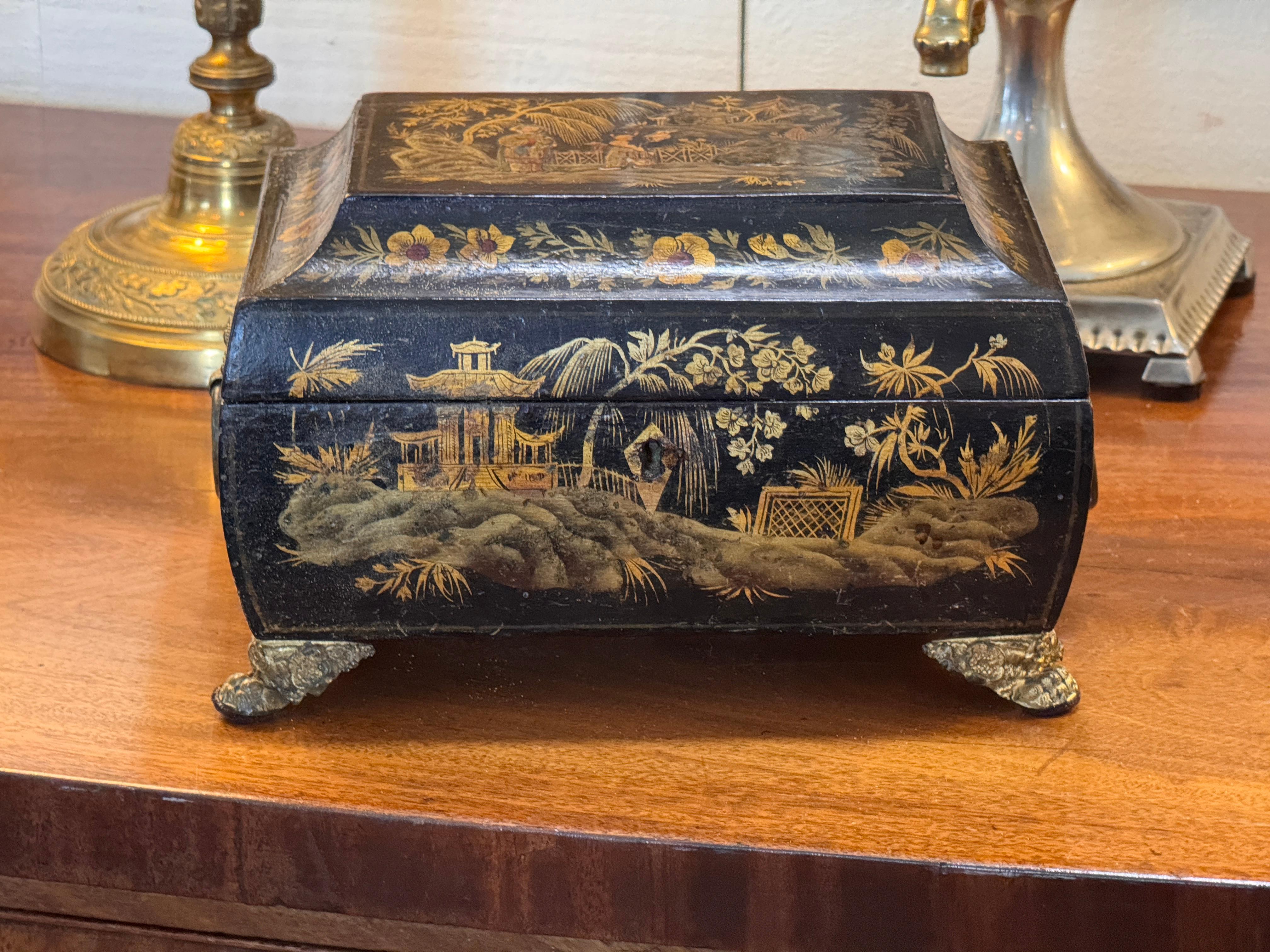 Chinoiserie box with hand-painted garden and pavilion scenes on all sides, including floral borders and a central landscape panel on the lid. The shaped body stands on cast metal feet, and the front is fitted with a decorative ring pull. Interior