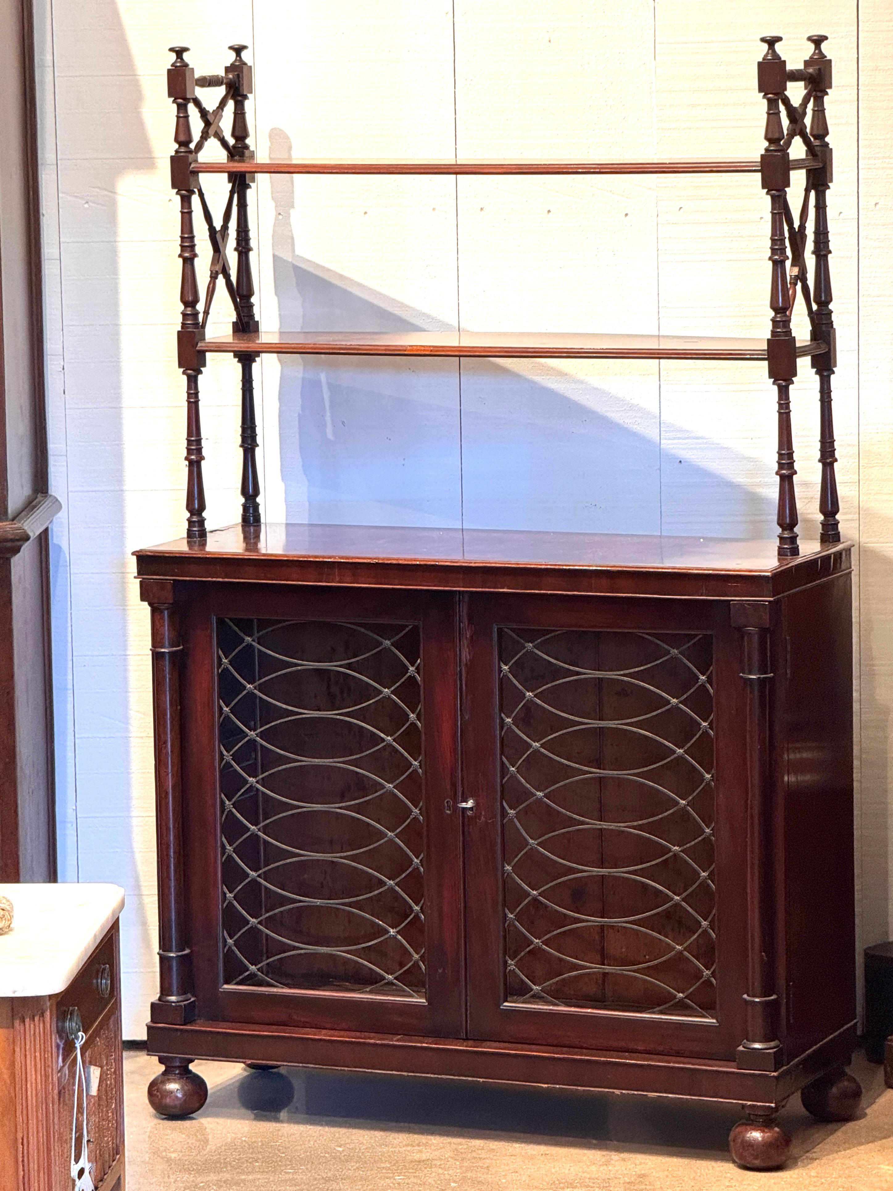 A 19th century Regency mahogany cabinet with upper open shelves supported by turned and X-form supports above a two-door base. The lower cabinet features gilt-metal grille doors with fabric backing, resting on turned feet. Rich grain and refined