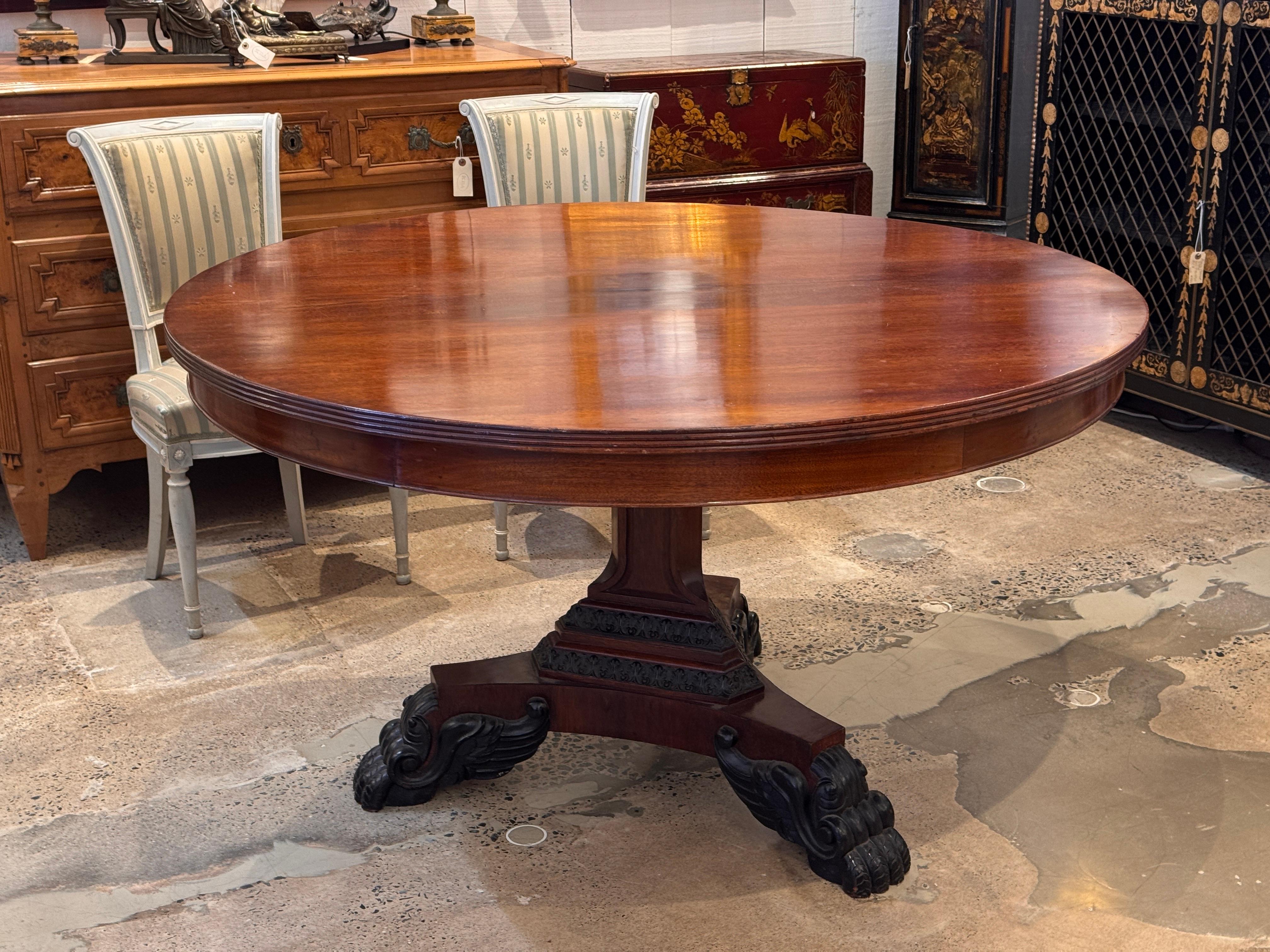 19th century Regency mahogany center table with a round top over a faceted pedestal. The base features carved foliate bands and bold carved feet shown clearly in the detail image.
