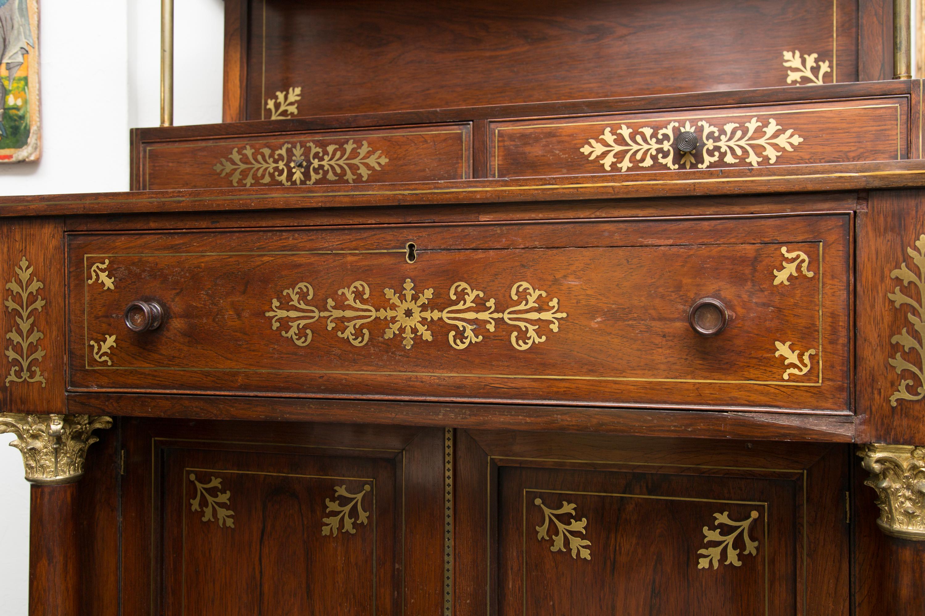 This is a beautifully articulated and unusual English Regency mahogany chiffonier secretaire. The top section containing two shelves with round brass supports over two small drawers with ornamental brass inlay. The top of the lower section is over a