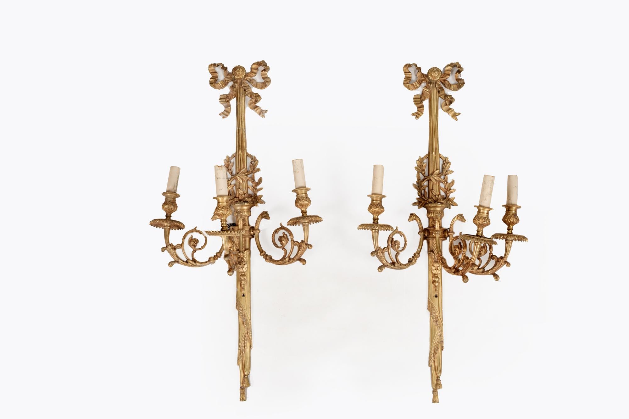 19th Century Regency pair of triple branch gilt metal wall sconces featuring elaborately scrolled candle holder light fittings with acanthus leaf and animal head detailing. The long stems are topped with cast ribbon details, the middle section