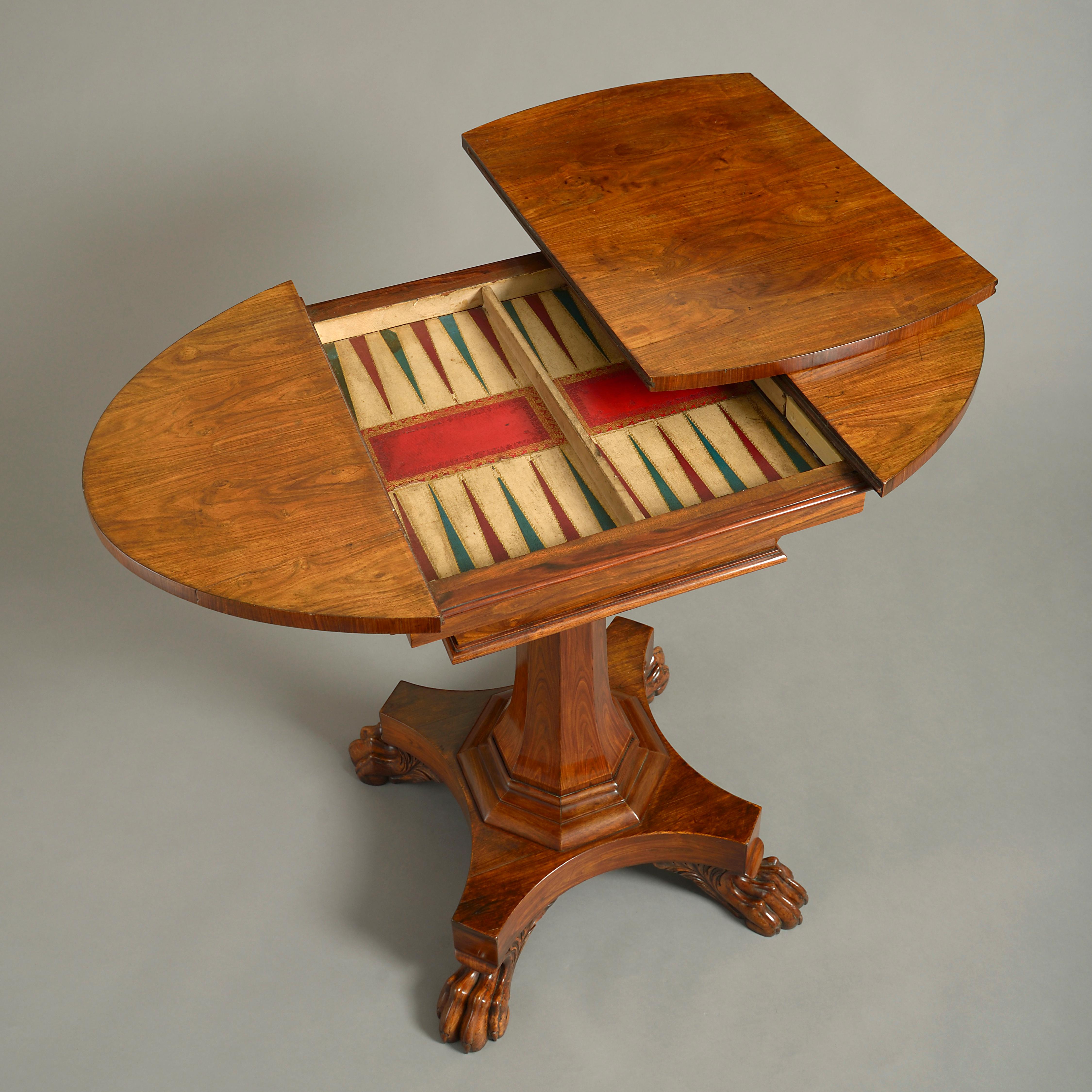 An early 19th century late Regency Period padouk wood games table, the oval top containing a recessed backgammon playing surface and raised upon a tapering octagonal stem, set upon a concave plinth, terminating in carved lion’s paw feet.