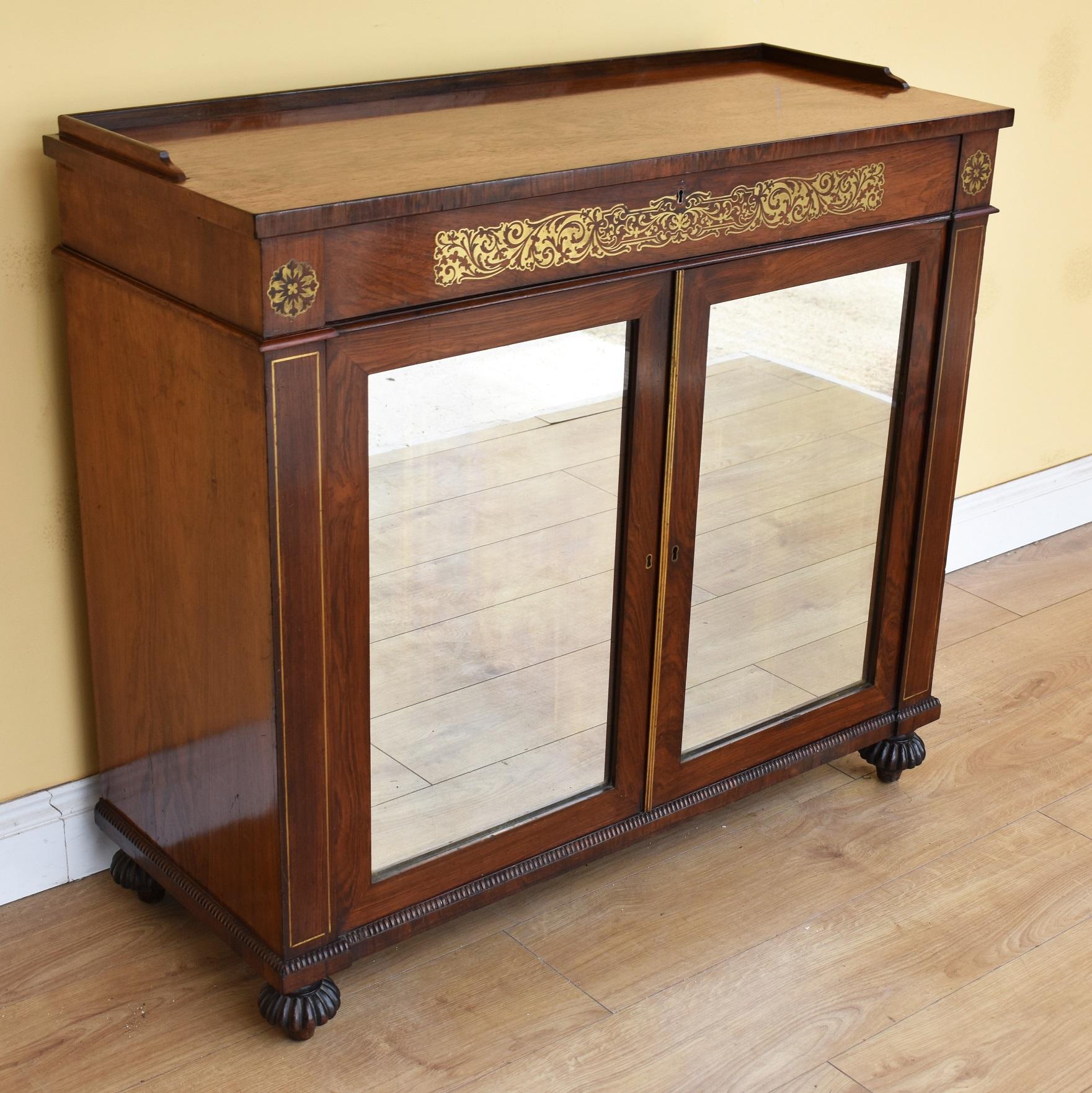 For sale is a good quality 19th century Regency rosewood and brass inlaid side cabinet, having a galleried top above a single drawer, decorated with ornate brass inlay, over two cupboards, each having mirrored panels, opening to reveal a single