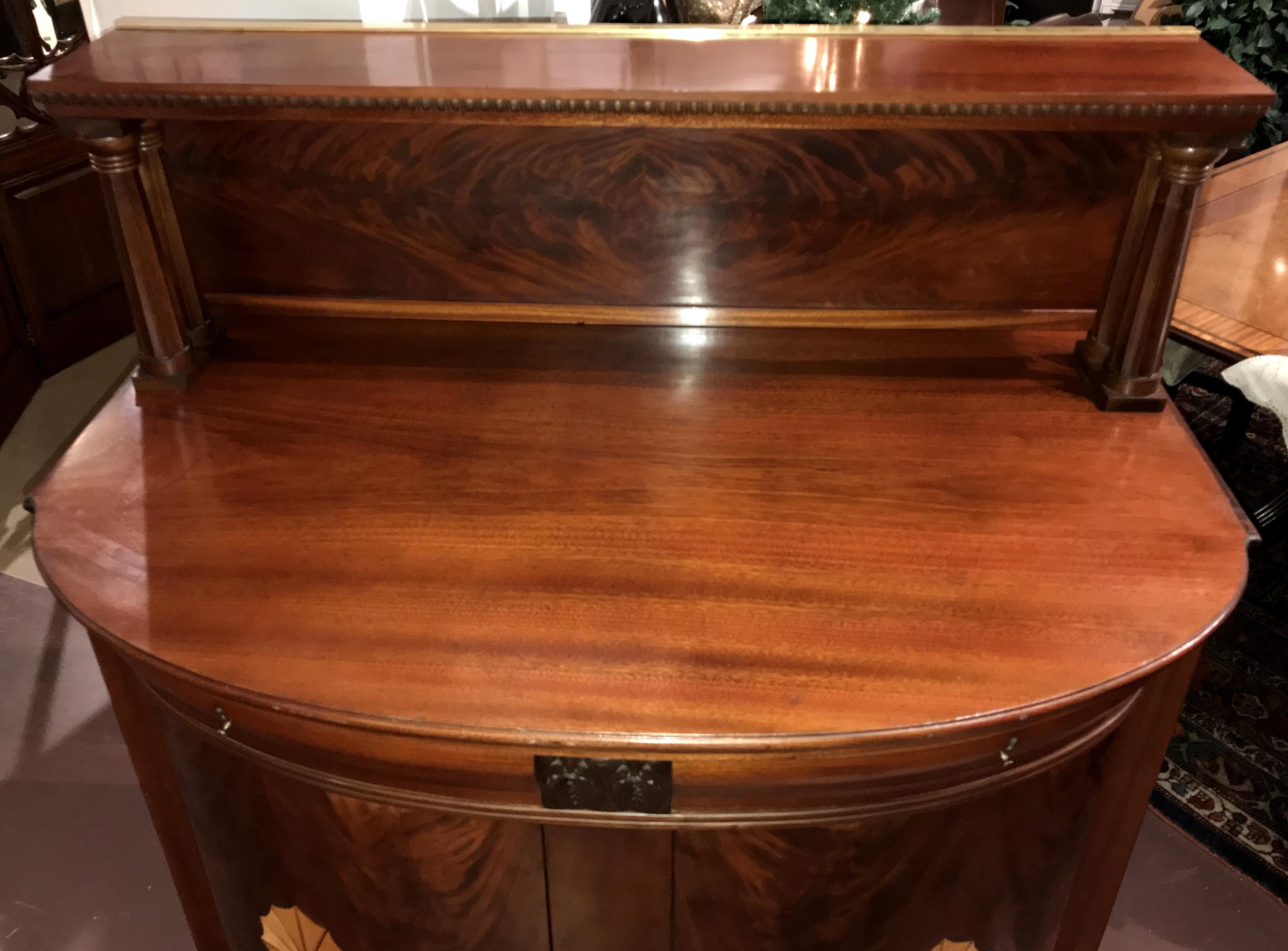 A wonderful English Regency style sea captain’s demilune form bar or server with raised shelf with backboard supported by two carved reeded columns on each side over a main case with molded frieze drawers, over two conforming cabinet doors, opening