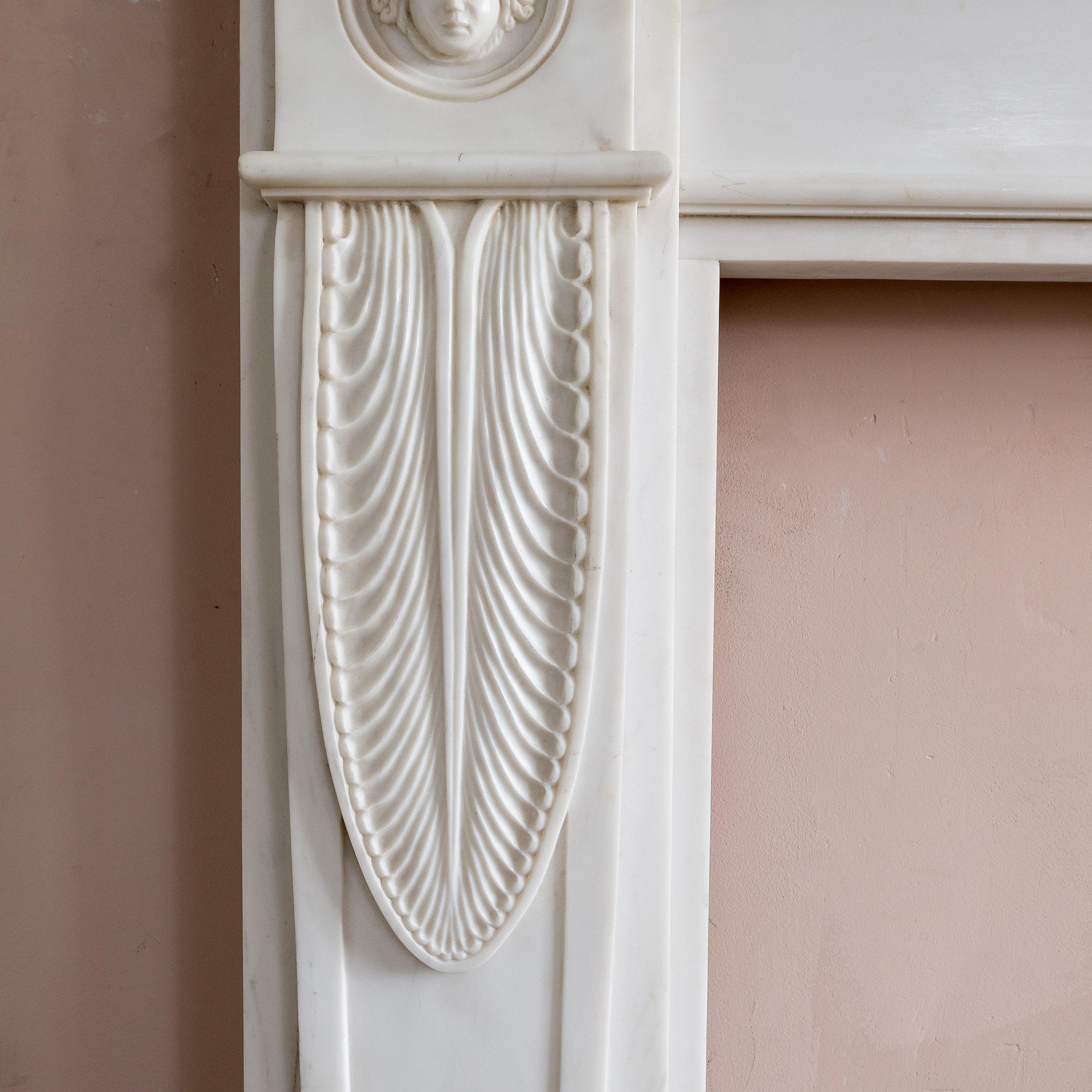 An early nineteenth century style white marble fireplace, modern, in the Regency manner, the frieze with plain, framed central tablet, the corner blocks with female masks set within circular frames above tapered jambs with delicately carved lotus