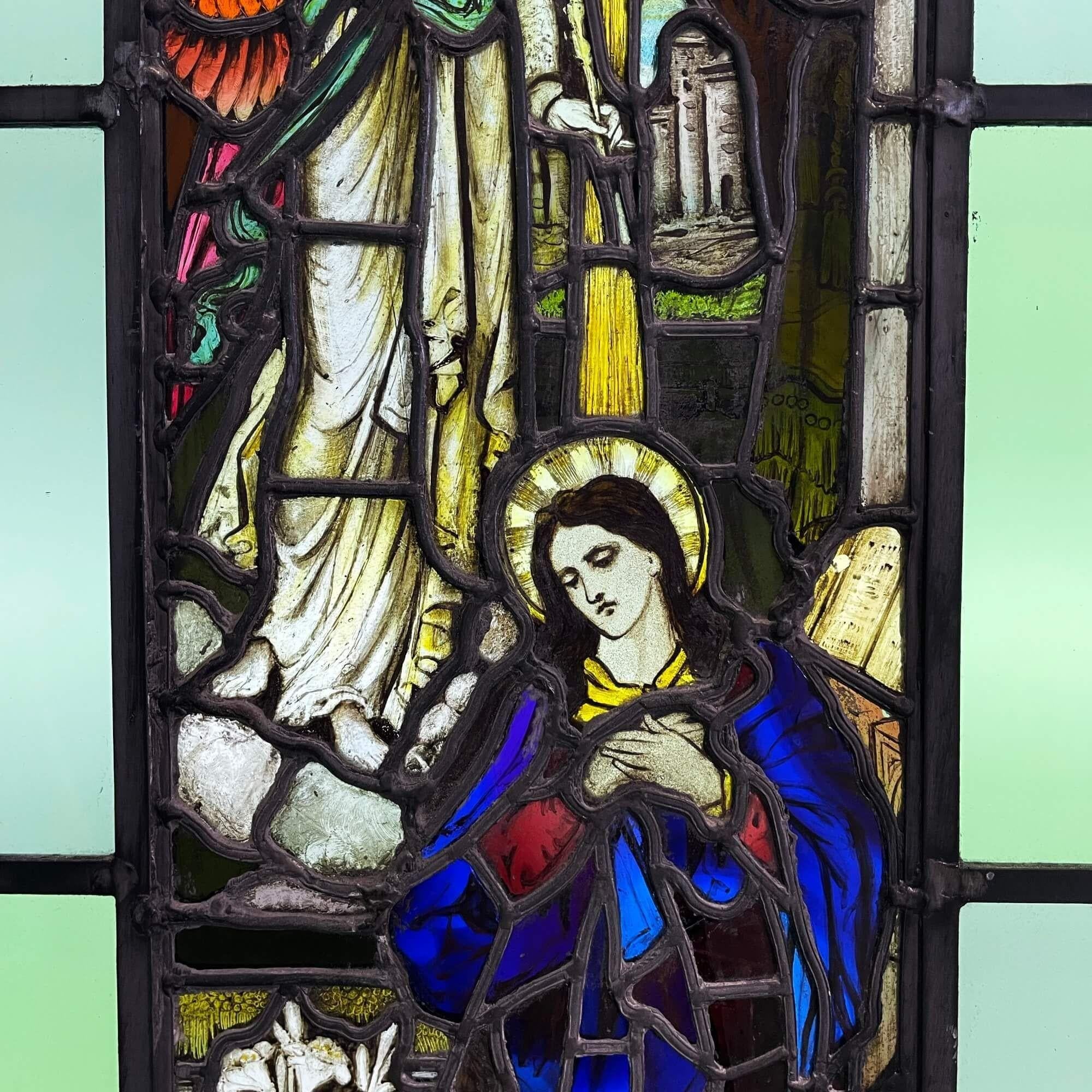 A mid 19th century religious stained glass window depicting the Annunciation, circa 1850. This vibrant stained glass window centres around a detailed panel illustrating the archangel Gabriel and Mary, dressed in blue. This is bordered by simpler