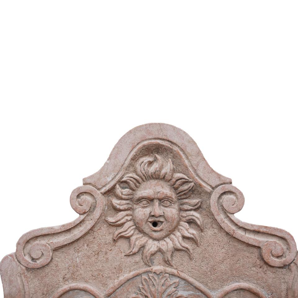 Ein antiker italienischer Wandbrunnen aus dem späten 19. Jahrhundert mit einer reich verzierten Aufkantung, die eine Sonnenmaske und Schriftrollen darstellt, auf einem schweren Becken mit seitlichen Schilden, das von einem verzierten Rollensockel