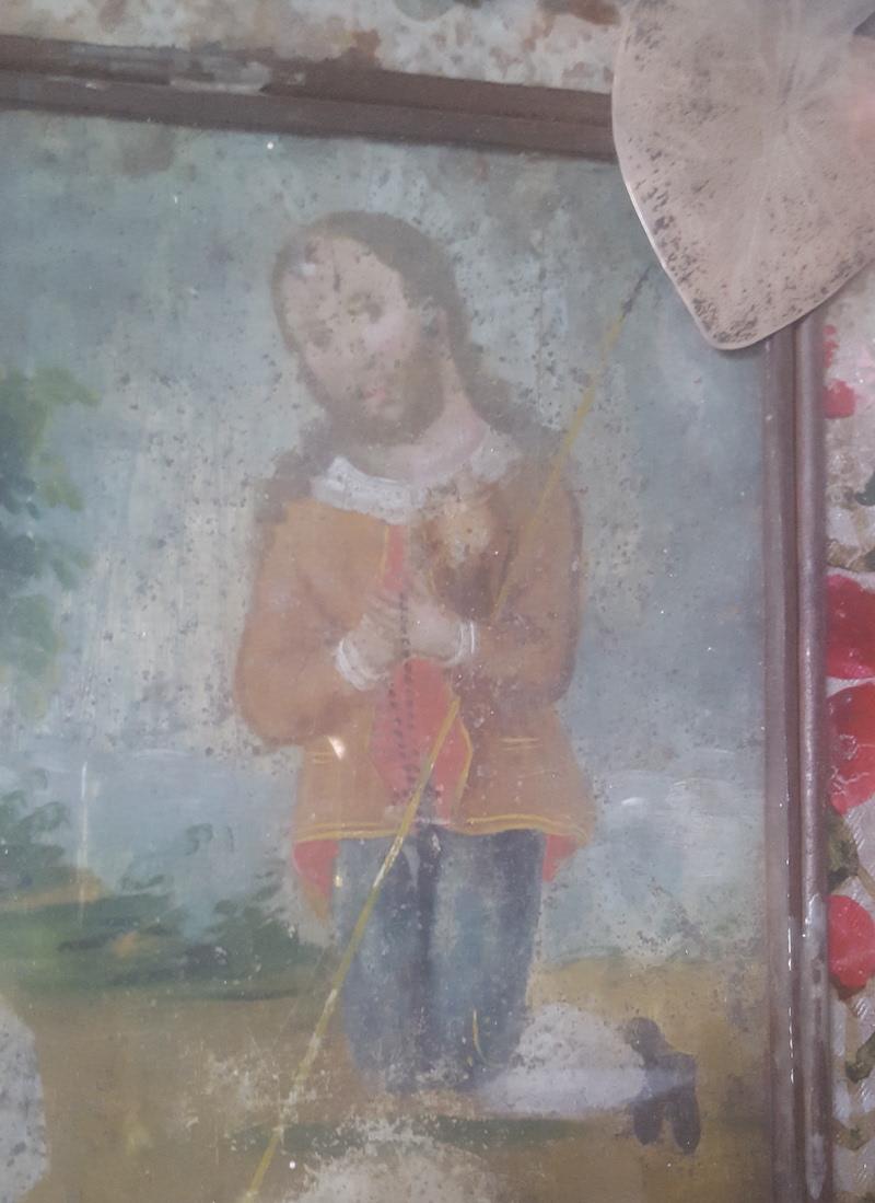 This painted Retablo on tin is in a distressed tin frame, very likely original to the piece. The tin edged frame encases glass over painted foil. The left side vertical, of tin, glass and foil is missing, while on the other intact side, a large,