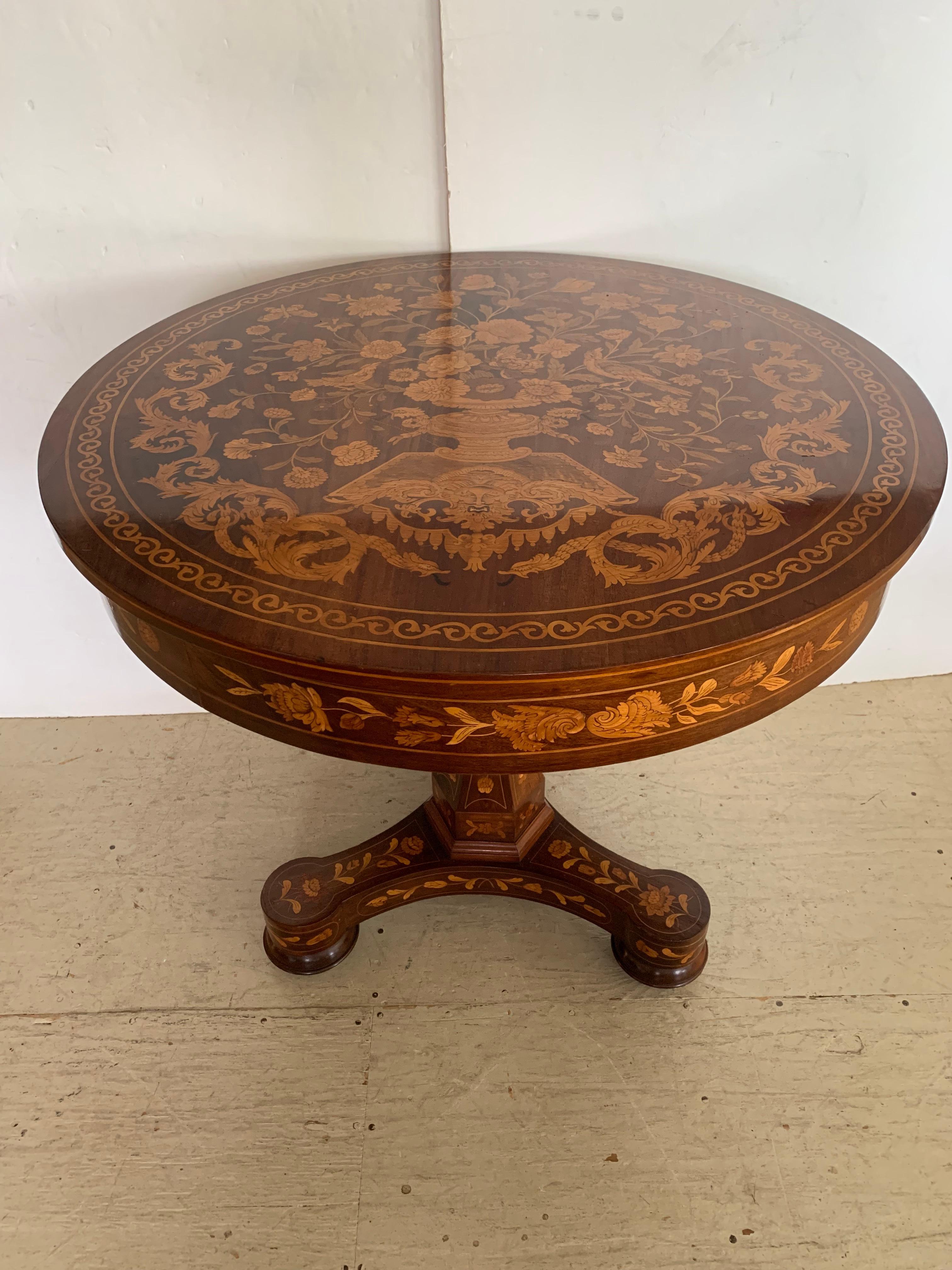 A marvelously decorative round foyer table or side table with elaborate inlaid marquetry on top, apron and base having floral and fauna and a central classical urn with overflowing bouquet. The faceted pedestal sits on a tripod base resting on bun