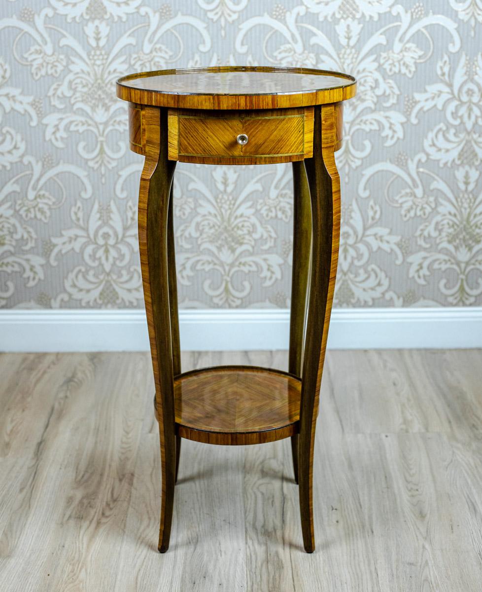 We present you a medium-sized end table/flower stand from the late 19th century.
The tabletop is round; with a single drawer underneath it.
This piece of furniture is supported on slender, slightly bent legs, which are joint together with a round