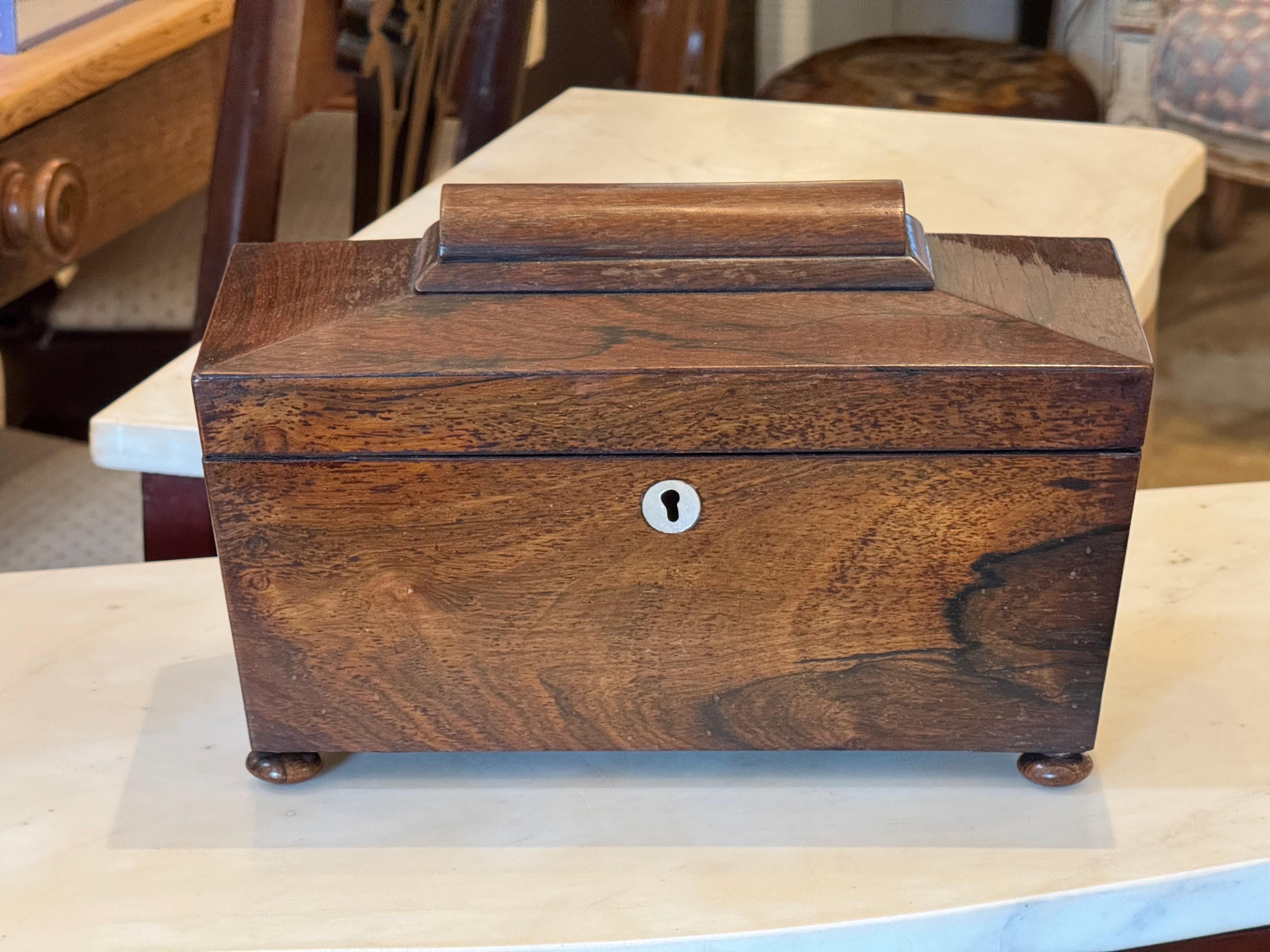 Teedose aus Palisanderholz mit geformtem Deckel und Stollenfüßen. Das Innere besteht aus zwei Fächern mit Deckel und einem Mittelteil mit einem Becher aus geschliffenem Glas, der wahrscheinlich zum Mischen verwendet wurde. Die Unterseite des Deckels