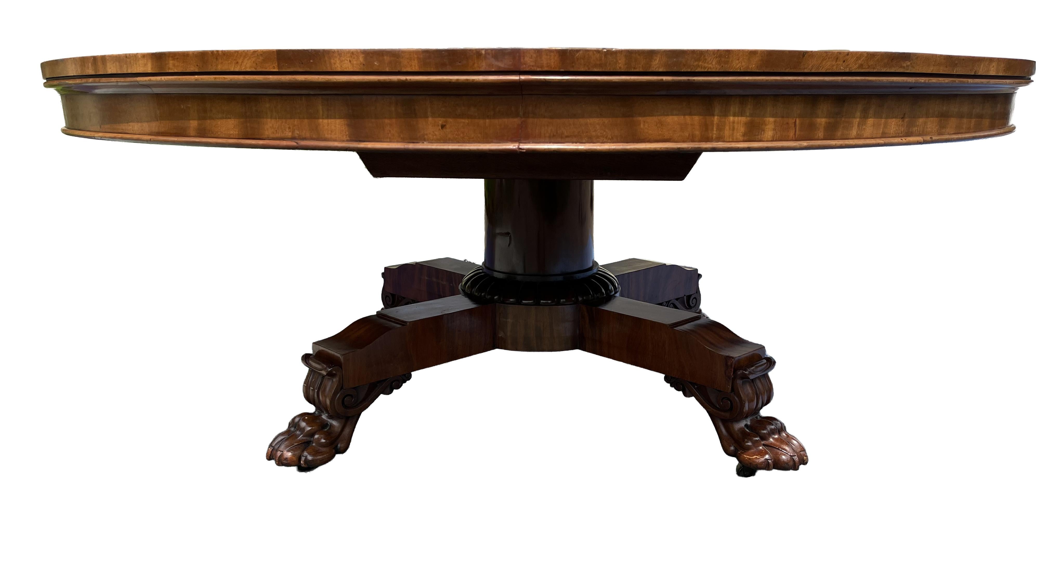A magnificent Regency-era mahogany round dining table boasts a vibrant color. The table features a top composed of two large boards with an apron. The tilt-top design is set above a substantial column with an intricately carved base. Four