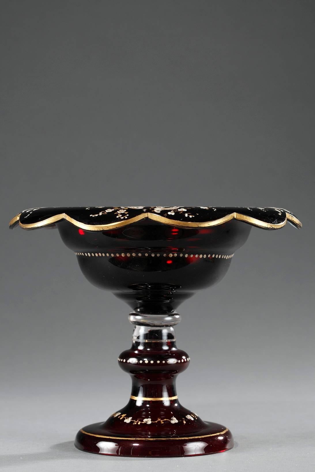 Ruby red, Bohemian crystal cup with a large, petal-shaped rim. It is decorated with enameled rinceau that is highlighted with gilded, flowering branches. The rim, stem and foot have gilded bands, and a ring of dots encircles the body of the cup and