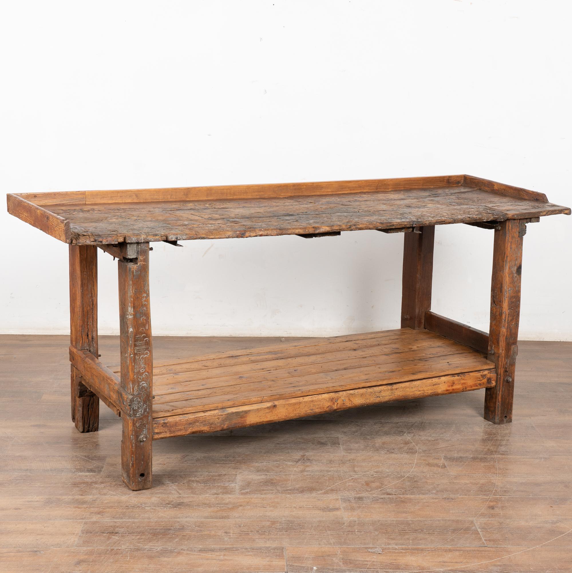 19th-century rustic pine work table with heavy distress and exposed bolts enhancing its industrial appeal. The surface shows extensive use with cracks, stains, gouges, and wear consistent with age. Lower shelf may have been replaced at some point in