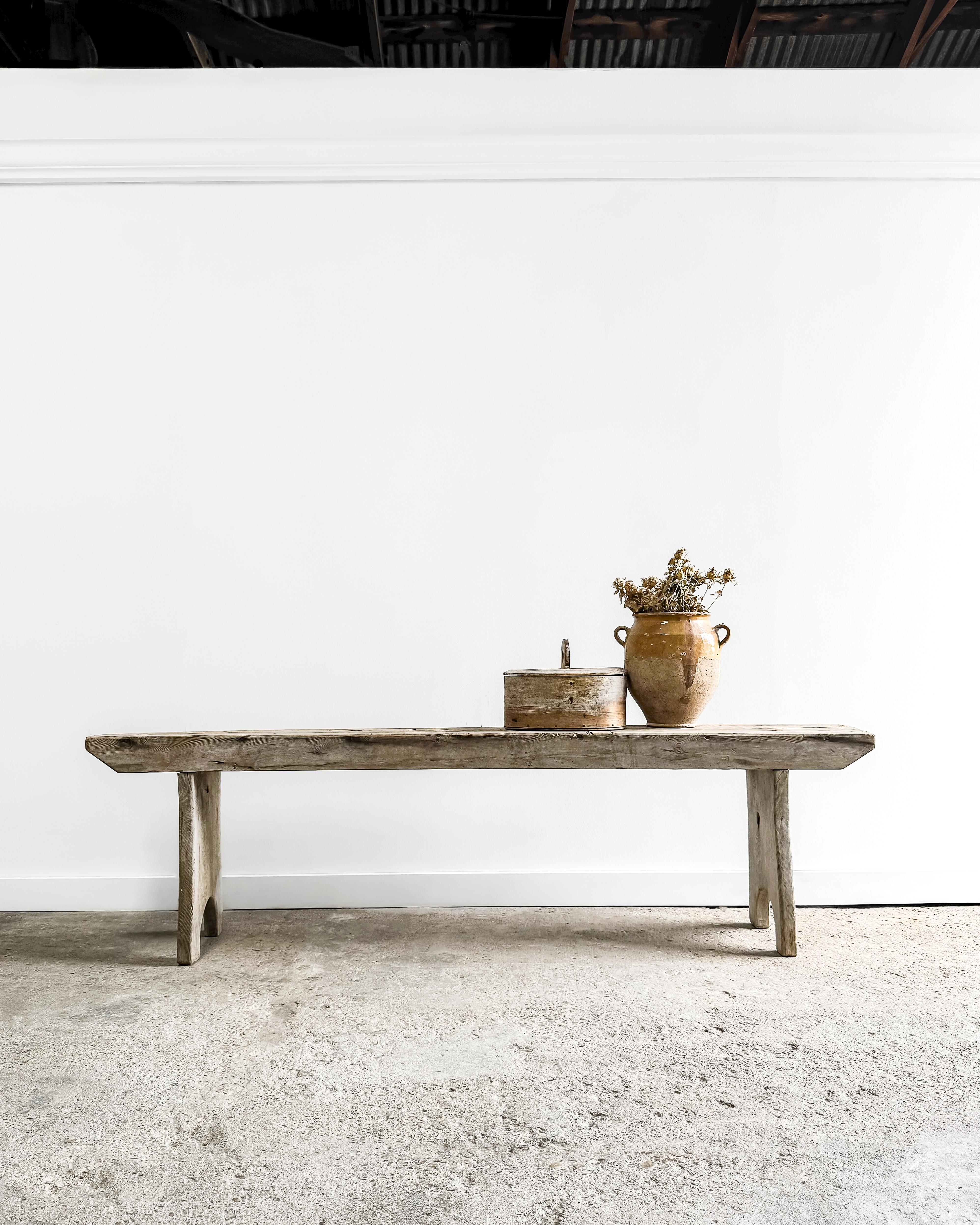 This hand-crafted white oak bench, featuring gracefully arched A-frame legs and a time-worn patina, embodies rustic charm and enduring craftsmanship. Its versatile design makes it an inviting addition to a large entryway, a cozy spot at the foot of