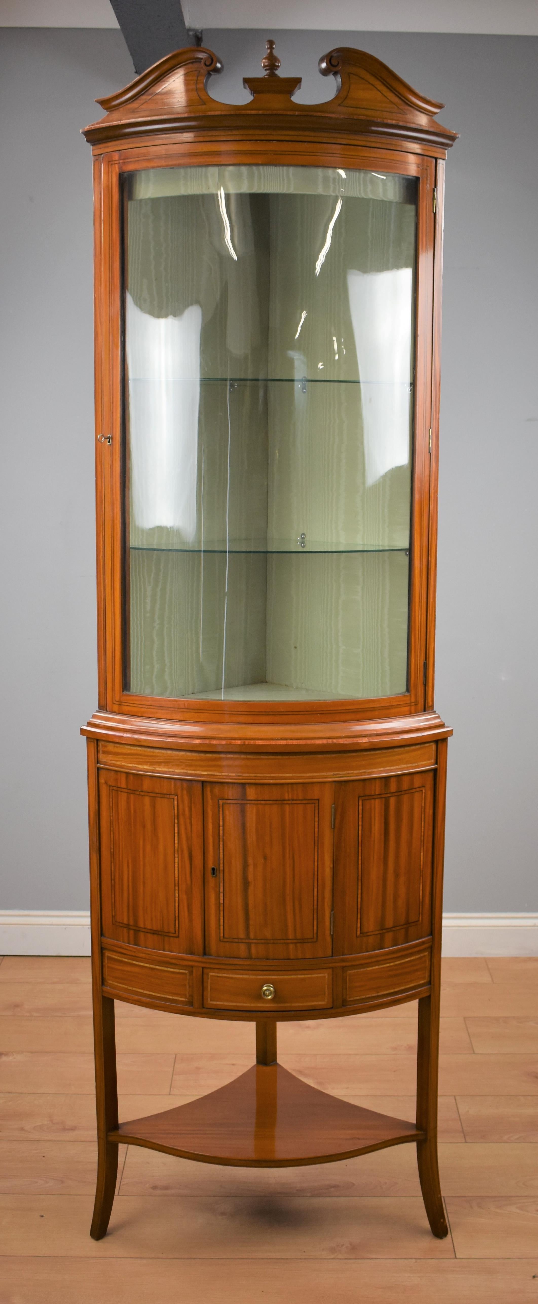 A 19th century satinwood corner cabinet display cabinet vitrine having a scrolled pediment top with bowed glass cabinet door raised over door and drawer base. Panels inlaid with boxwood. Raised on shaped saber legs united by tier stretcher.