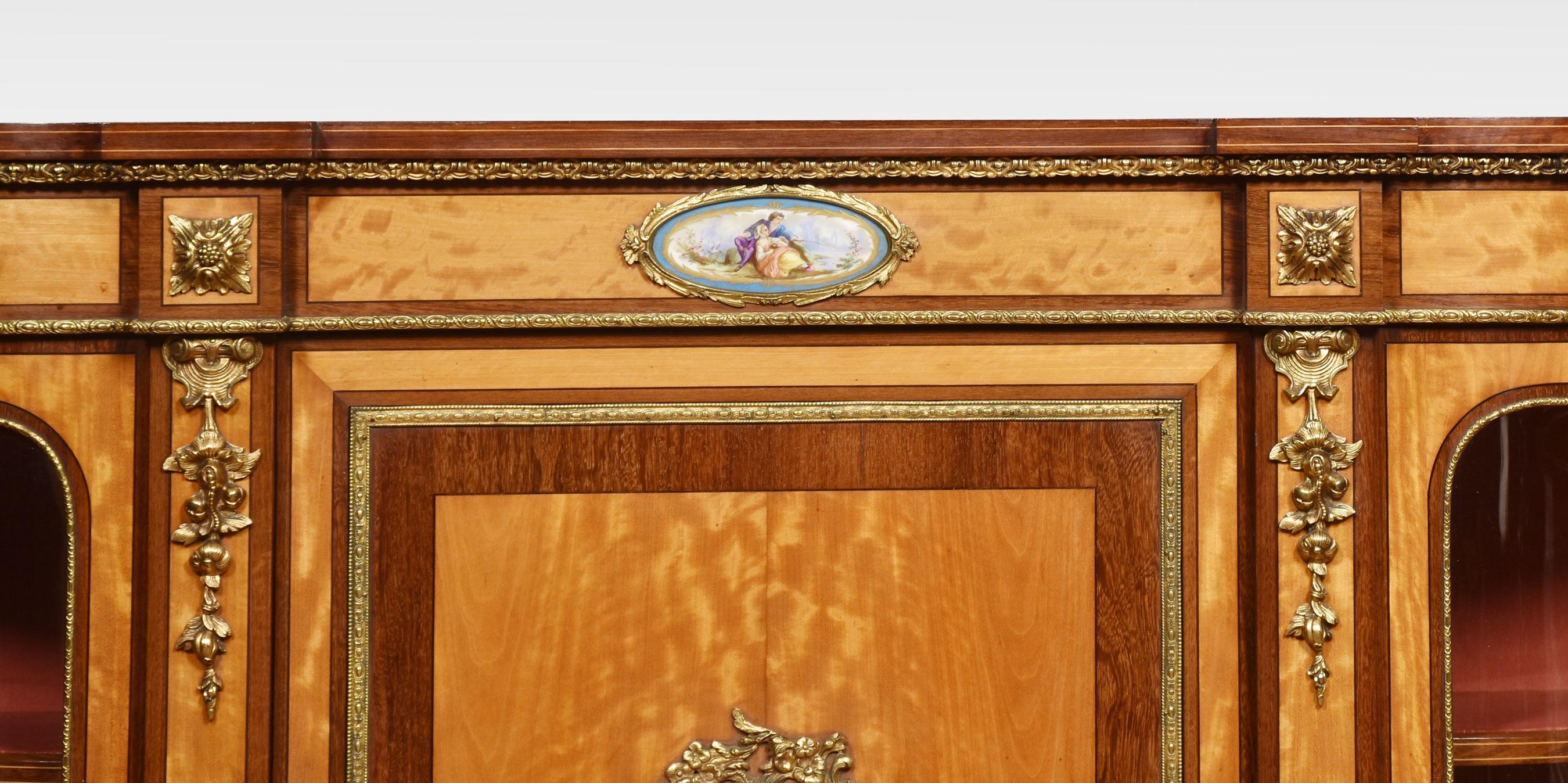 Brass mounted satinwood credenza, the well figured top having a crossbanded edge. Above a central door with Sevres porcelain oval panel. The ends fitted with bowed glazed doors enclosing velvet-lined shelves. All raised up on short bun