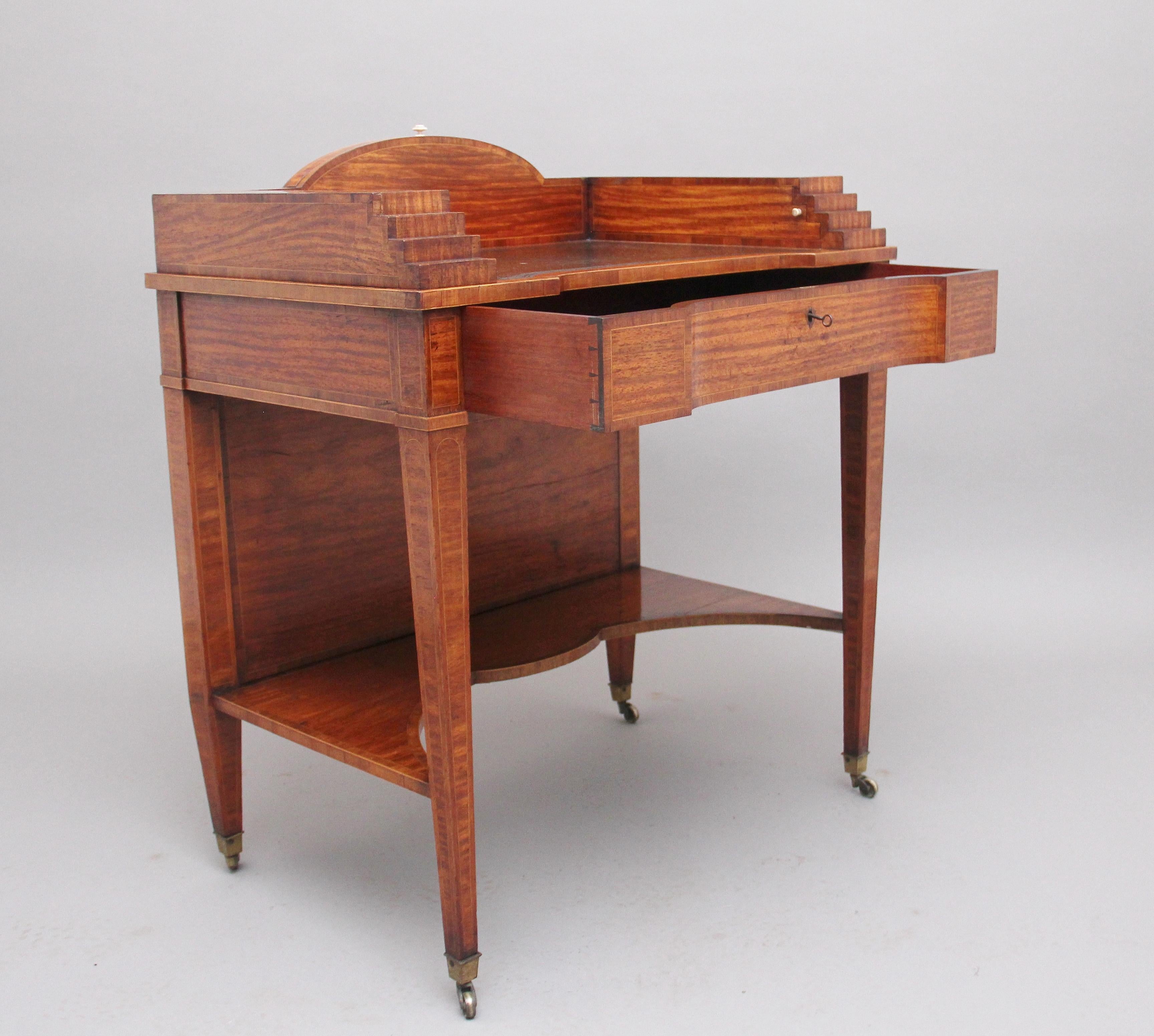 19th century satinwood ladies writing table in the Sheraton style, having a shaped leather writing surface decorated with blind tooling, decorative stepped sides which has secret pull out compartments either side to reveal a pen tray on one side and