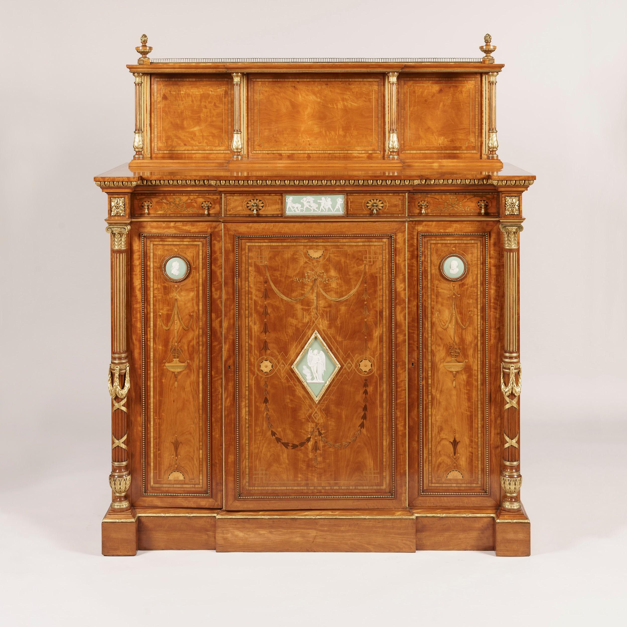 A fine side cabinet
firmly attributed to Wright and Mansfield.

Constructed in a brilliant satinwood, and skillfully incorporating tulipwood, harewood and various specimen wood marquetry work, and expertly carved giltwood accents; of gentle
