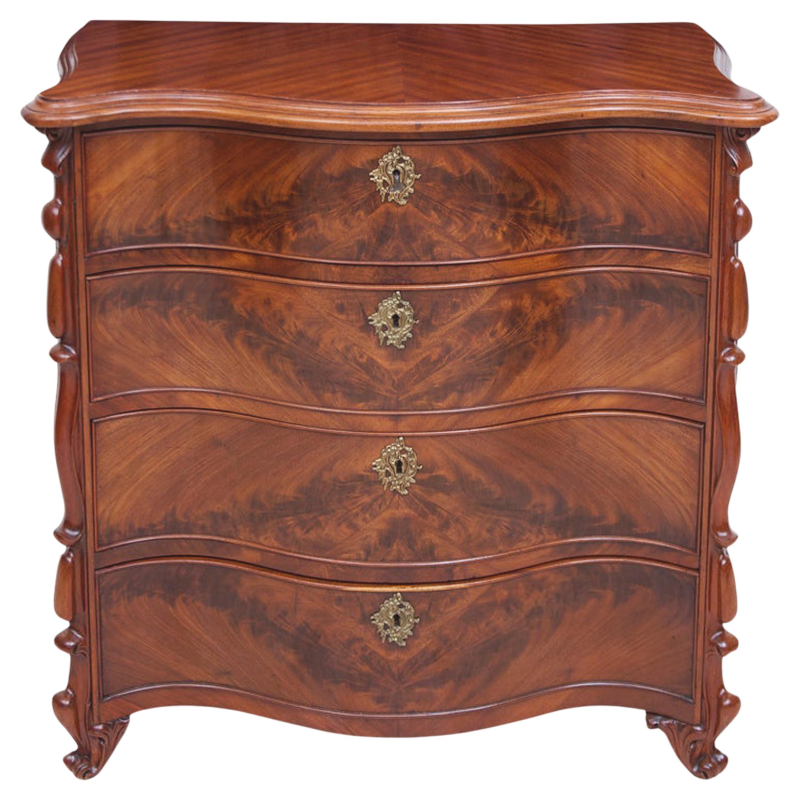 A Fine chest with four drawers in bookmatched, crotch West Indies mahogany with serpentine front and resting on carved slipper feet. Features a serpentine top in quarter-sawn sapele (African mahogany) with mahogany sliding drawer covers inside top