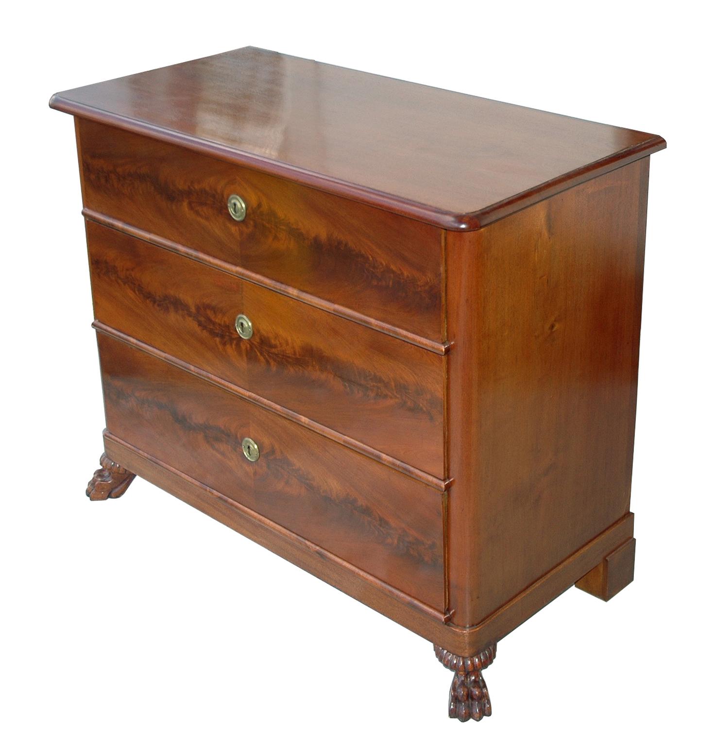 A handsome Empire commode made of fine West Indies (Cuban) mahogany with book-matched flame mahogany veneers on the drawer fronts. This chest offers three large storage drawers and rests on carved lion's paw feet. Restored with French-polish in our