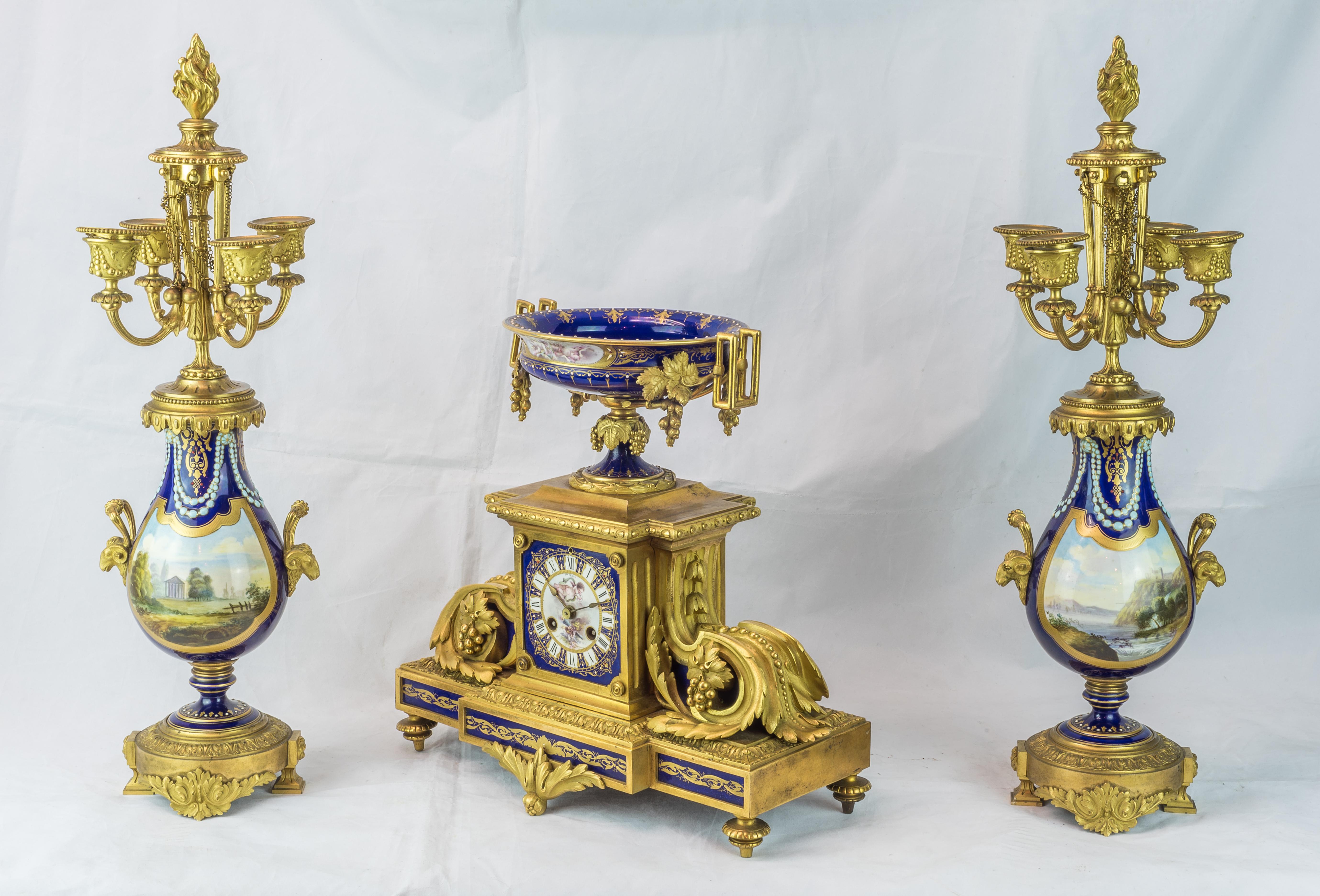 A magnificent Sèvres style gilt bronze and cobalt-blue 'Jeweled' painted porcelain clockset.
The central clock case surmounted by a ruby and pearl 'jeweled' coupe flanked by bracket handles; the vasi-form four-light candelabra with similar jewels