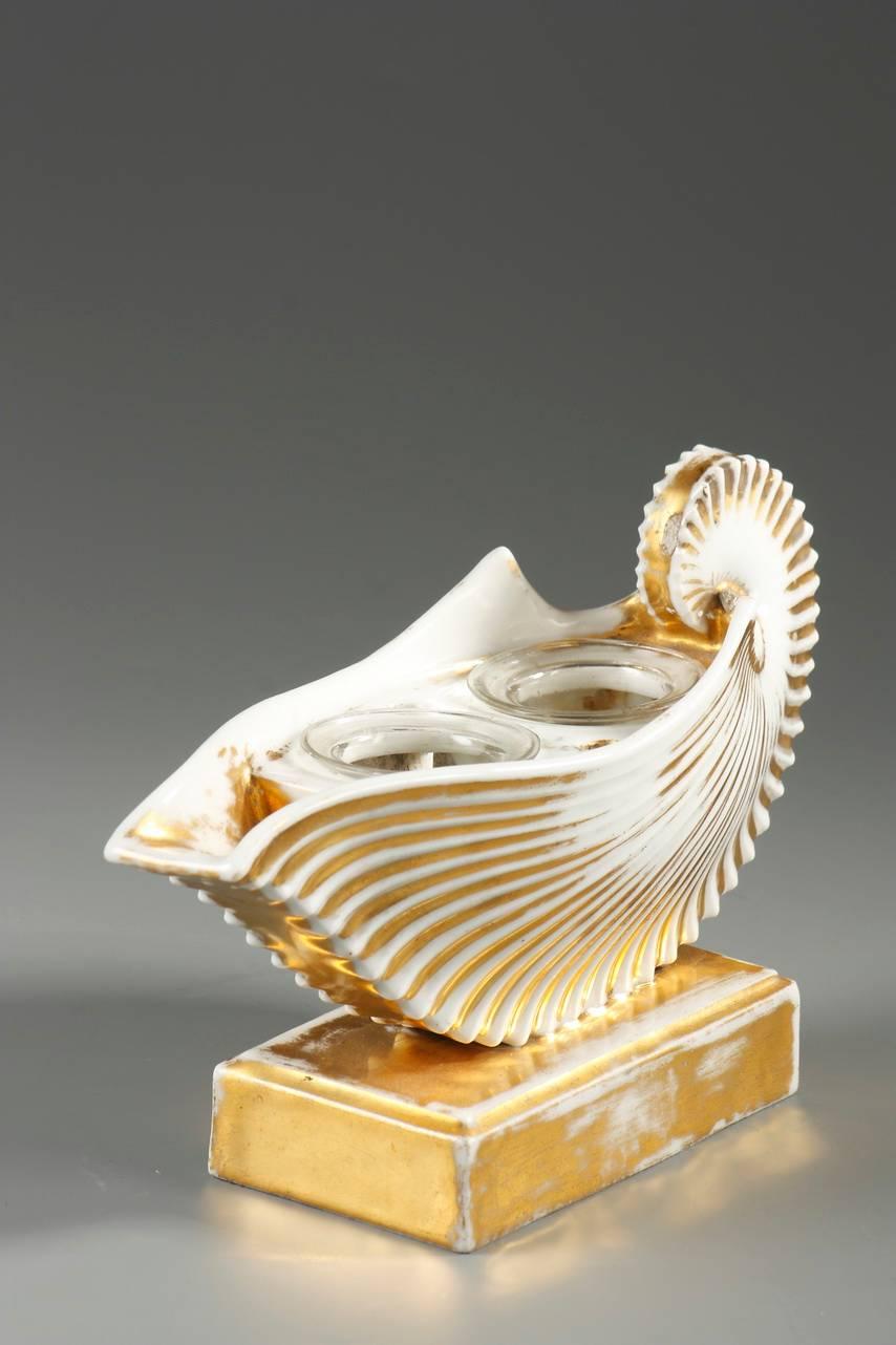 White porcelain inkwell in the form of a shell, highlighted with gold and resting on a square base. The top features two round, glass inkwells and two pen holes. Light wear to the gilding and light nick on the base,
 
circa 1820.

Dimensions: W
