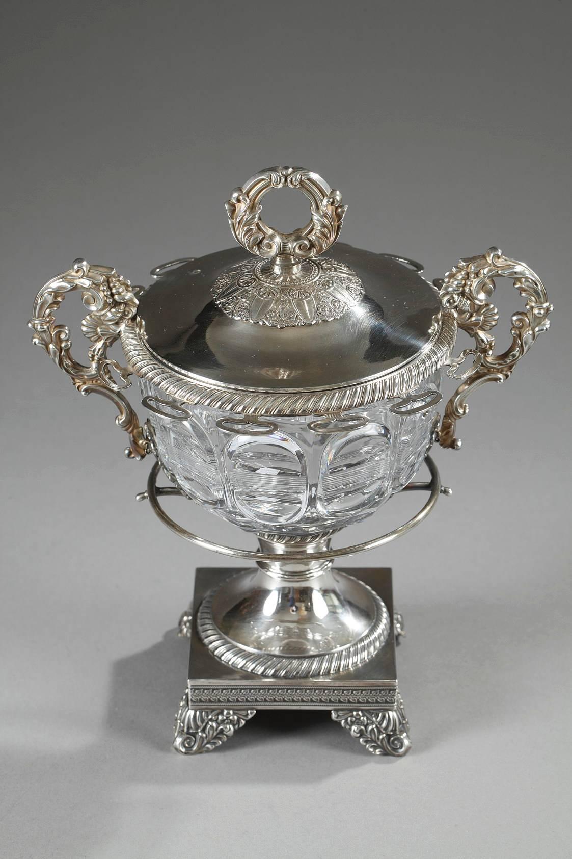 Silver and cut-crystal candy dish decorated with geometrical patterns. The two silver handles are richly adorned with acanthus leaves, scrolling foliage, shells, and palmettes. The removable lid is ringed with a spiralling frieze, and it is topped