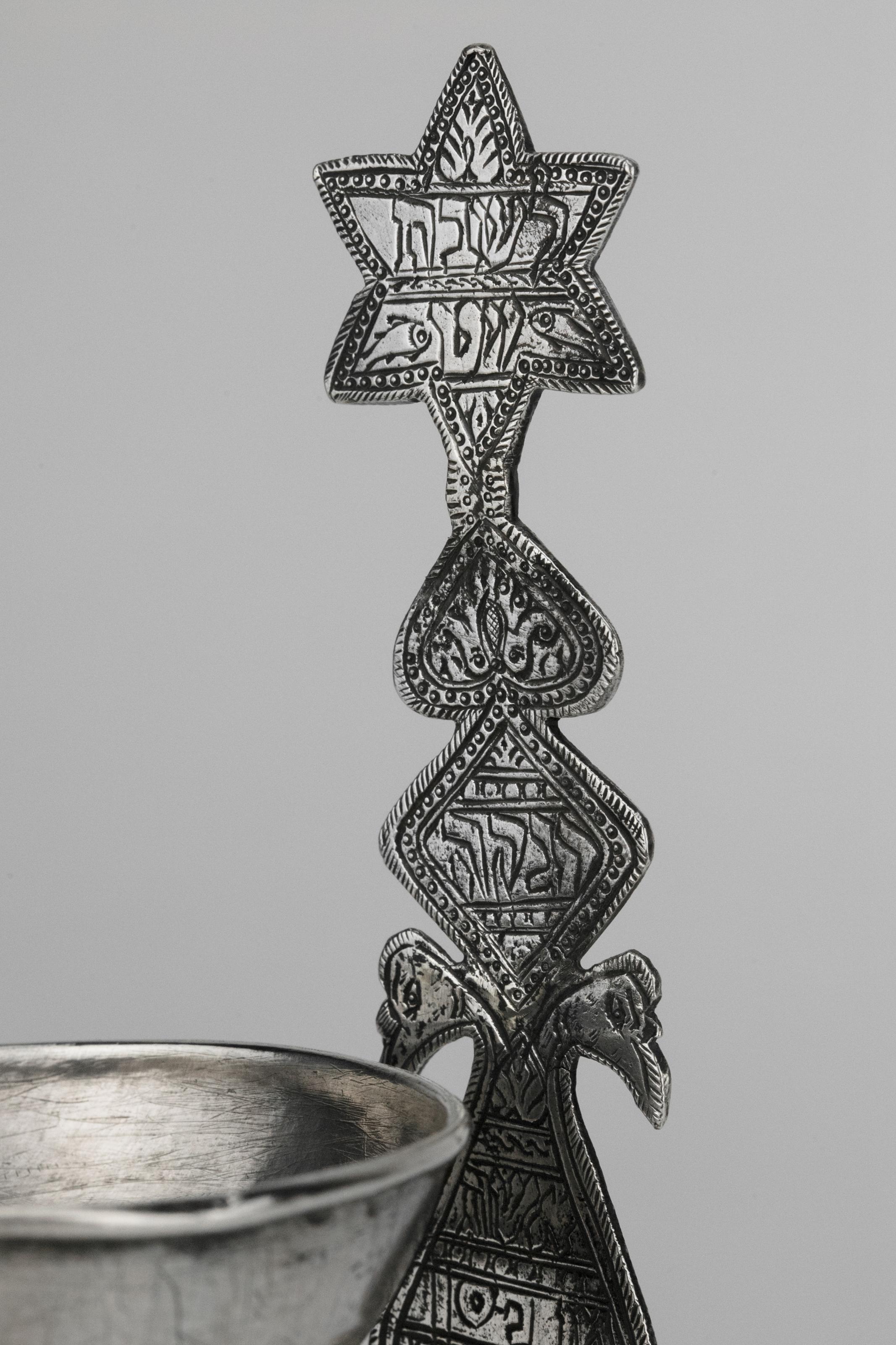 Silver Sabbath oil lamp, Afghanistan, late 19th century. 
Featuring a rimmed bowl elevated on a paneled stem, with larger drip pan below, resting on three feet. Extended piece of hand cut silver at rear, with Hebrew engraving inside the Star of