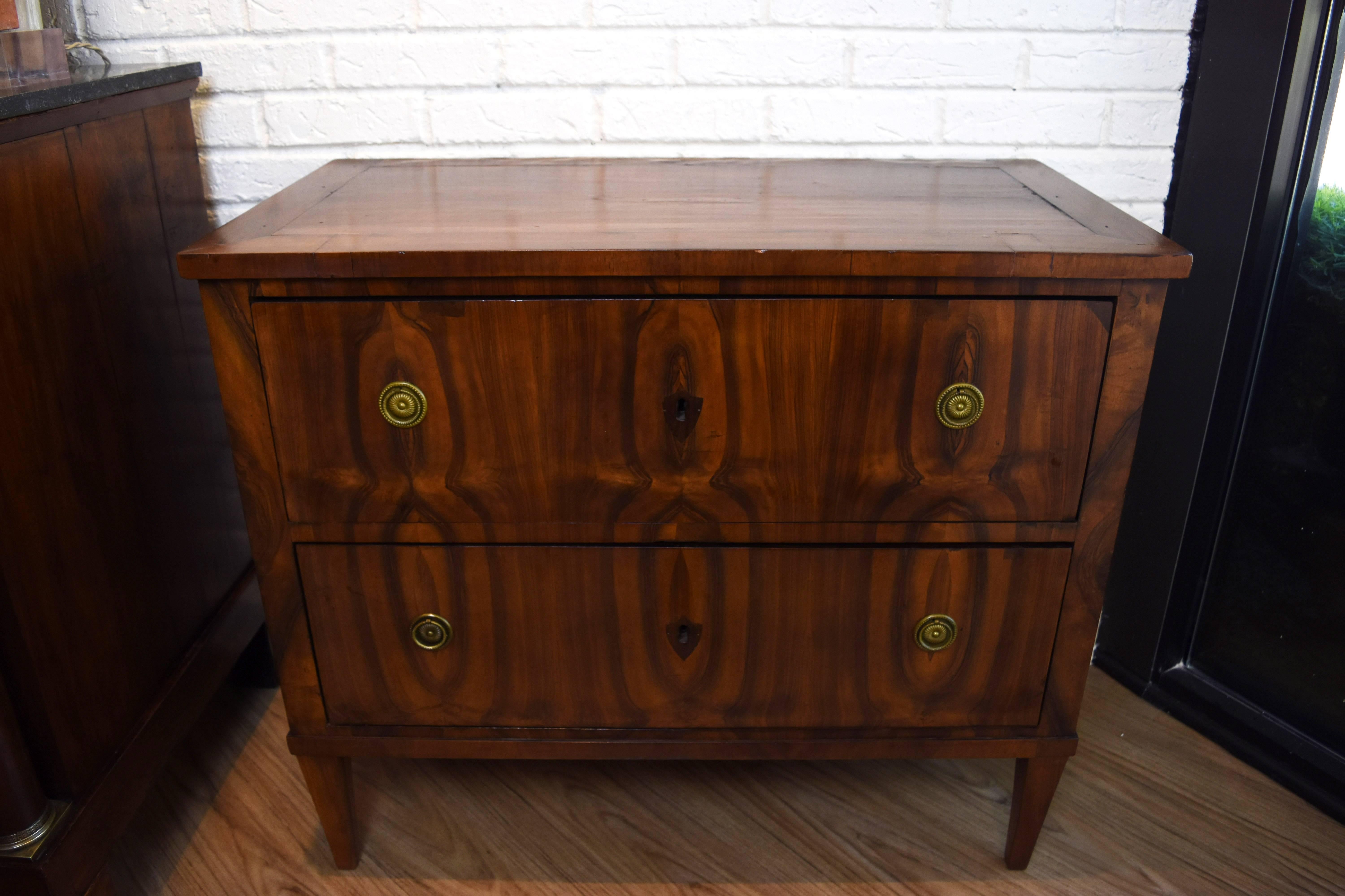 This Biedermeir commode features two drawers with book matched walnut and inlayed wood key escutcheons. The brass pulls are original and the legs are tapered. The size of this commode would make it perfect for a bed side chest.