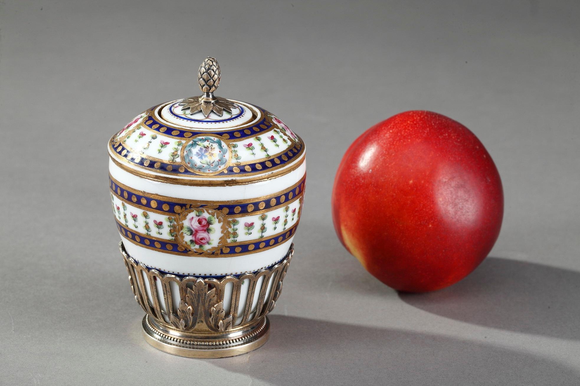 Small inkwell crafted in porcelain, the paunch and lid decorated with a frieze of polychromatic floral motives highlighted with gold. The lid topped by a pine cone finial features a compartment to hold ink. Silver-gilt mounts, finely chiselled with