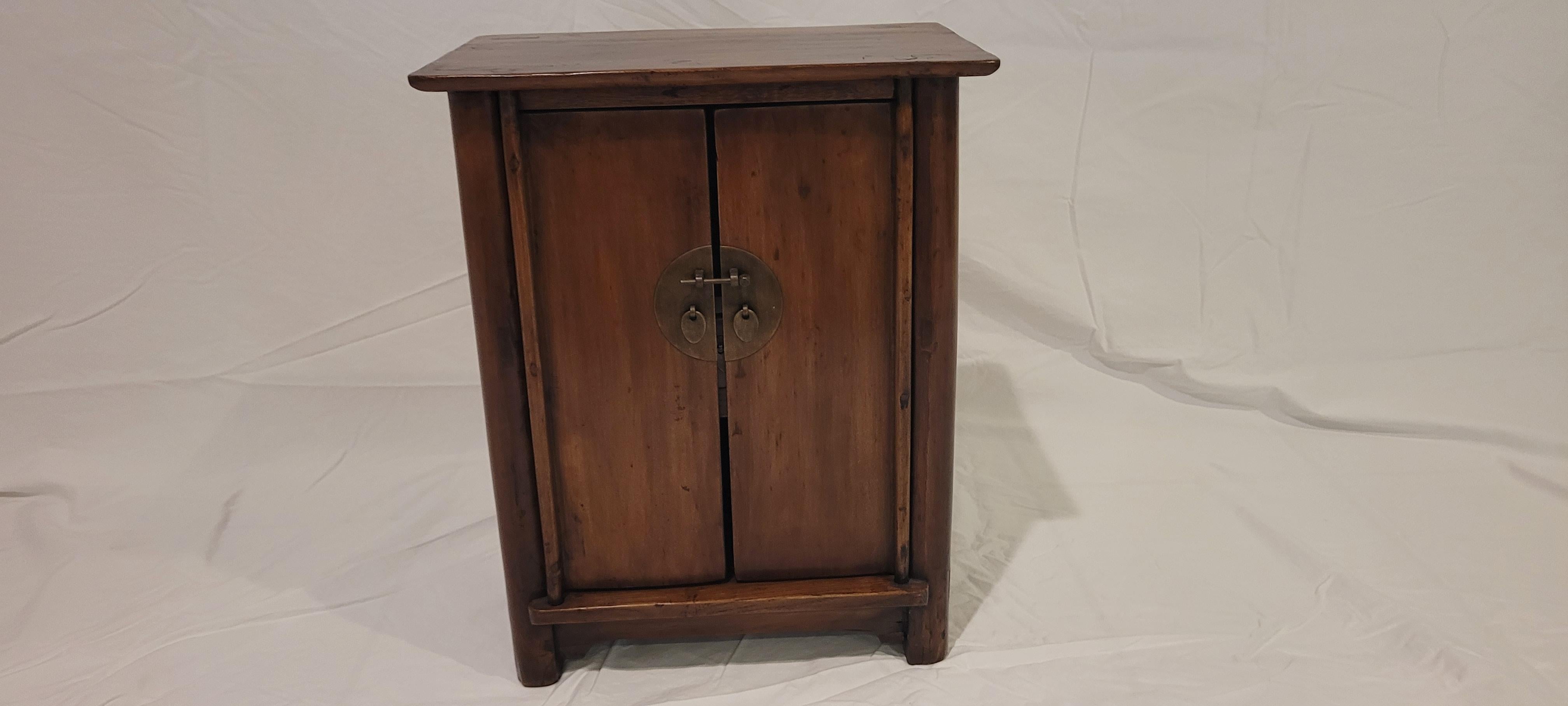 Small Kang Cabinet	18.5h x 14.75w x 10d
This small Kang cabinet has a flush top which slightly hangs over the body frame.  The frame members are round and the front is decorated with a round metal plate.  The apron is simple without any