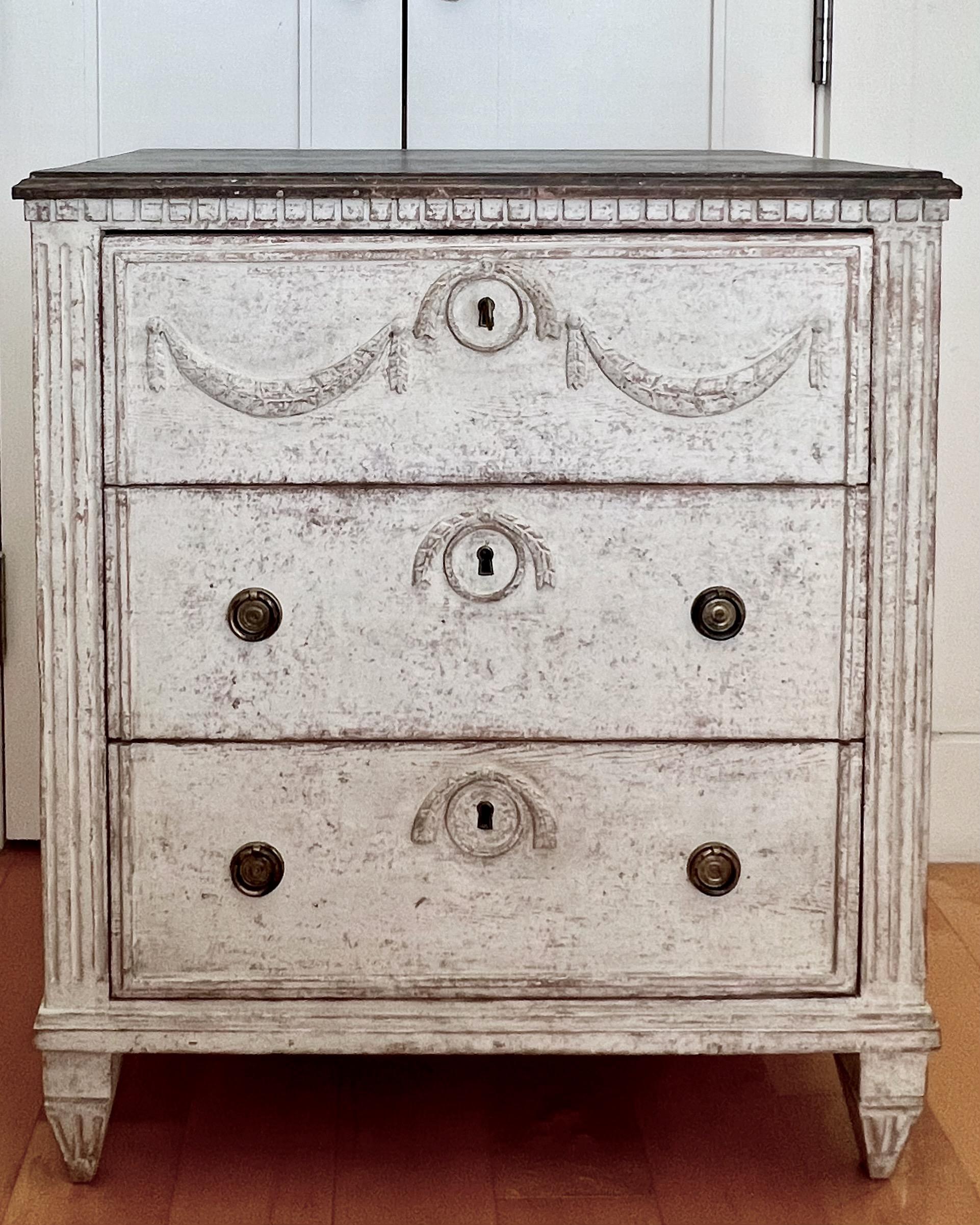 Charming 19th century Swedish three drawer chest with carved dentil moldings, canted corners, fluted side post and classic ribbon and swag decorations, painted in light gray with blackish wooden top.
Sweden 