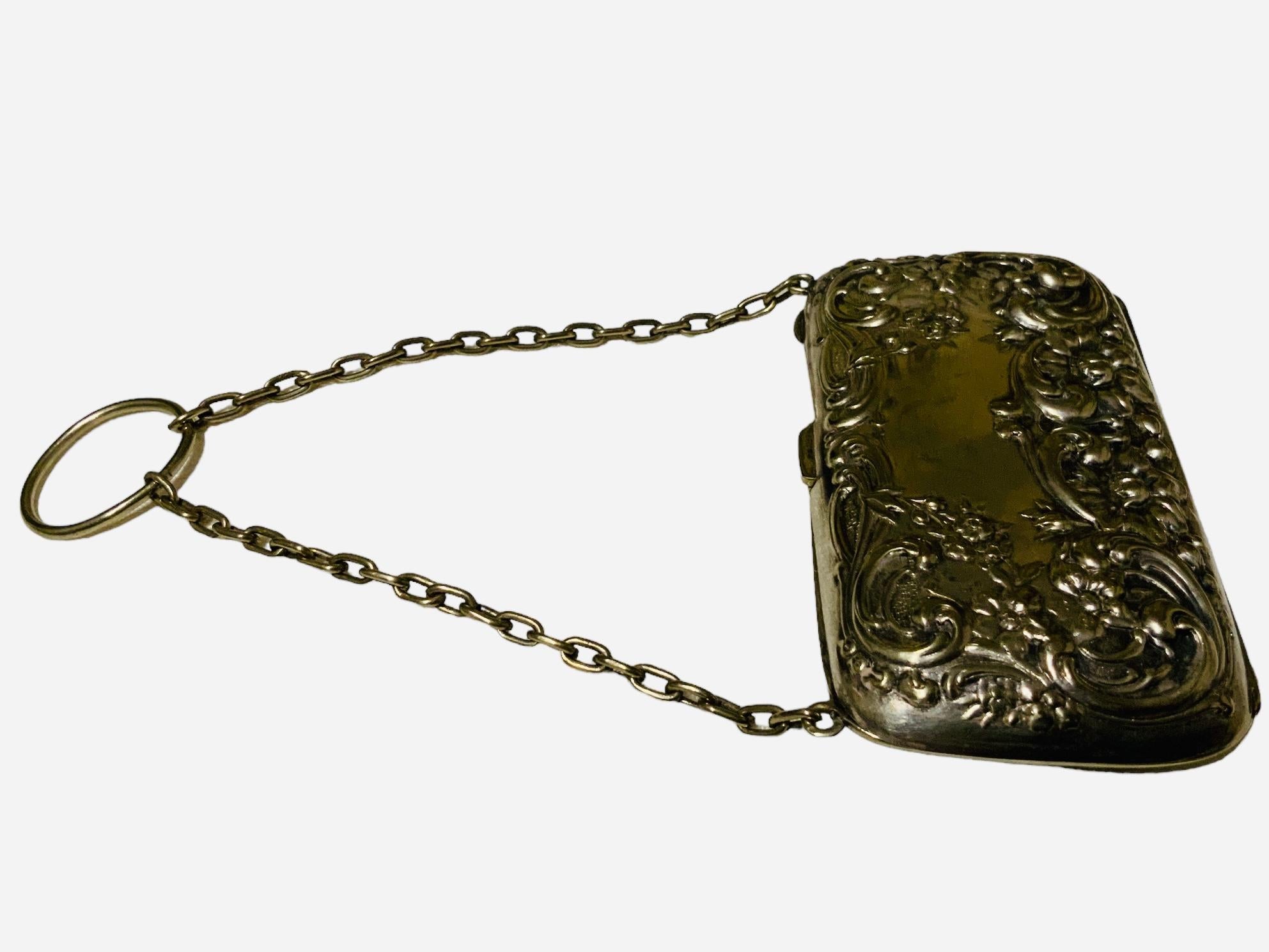 This is a Smith & Bartlam EPNS Finger Coin Purse. It depicts a rectangular electroplated non sterling silver purse decorated with a high relief of scrolls of acanthus leaves, small and large flowers in the front and back. Small silver chain with a