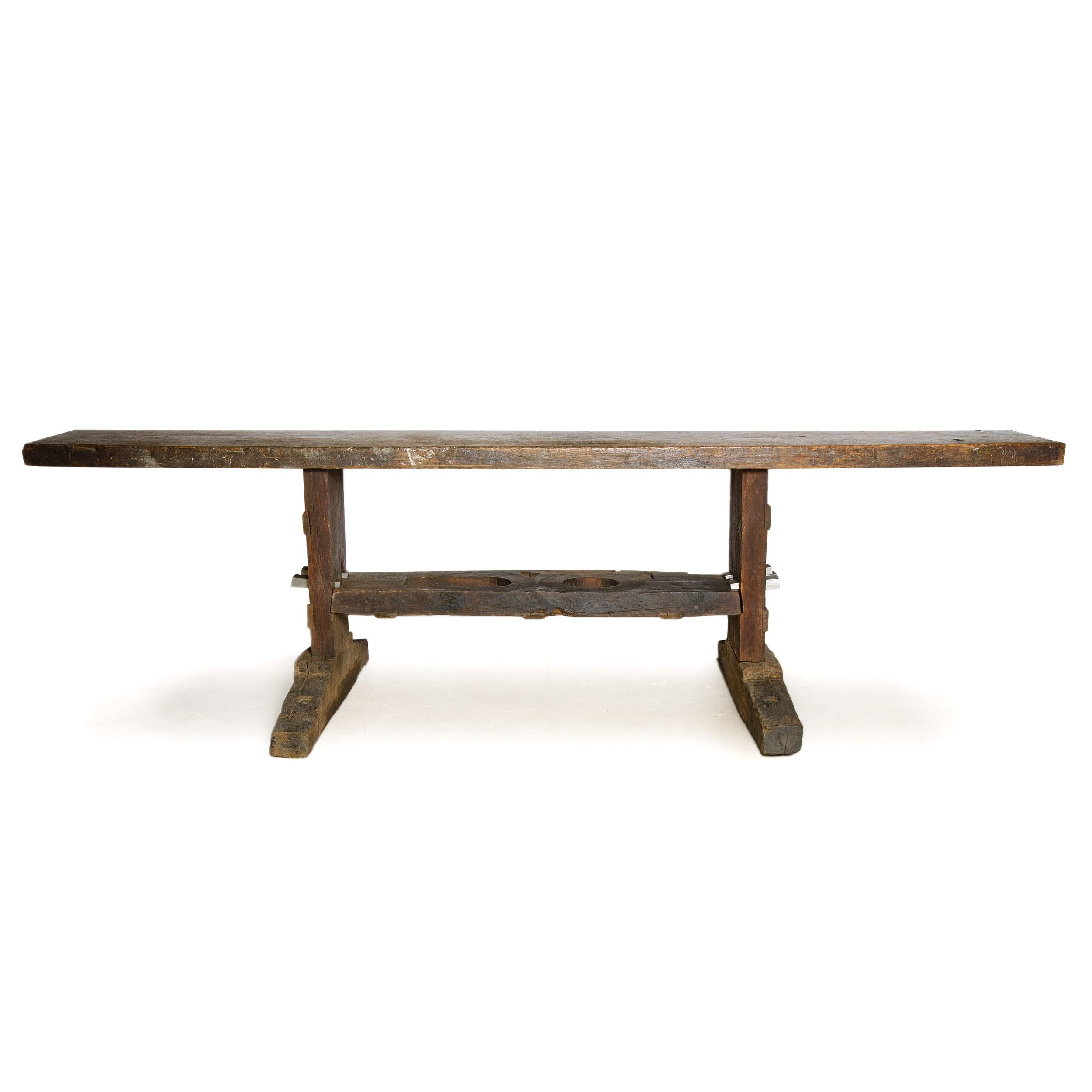 Importante banco de trabajo antiguo, de roble macizo, ejecutado en la forma tradicional de una mesa de caballete con pie de zapata, con su amplia repisa inferior como elemento de refuerzo espigado en ambos soportes laterales. No es una pieza