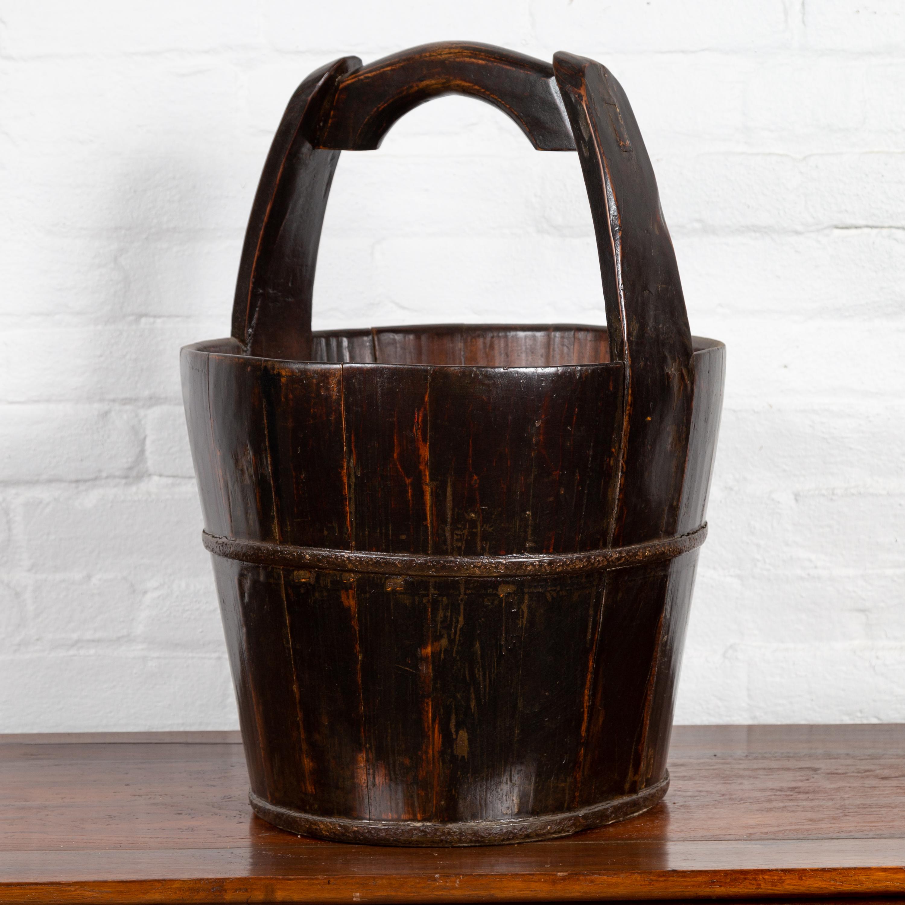 A Chinese rustic wooden bucket from the 19th century, with large curving handle. Born in Southern China during the 19th century, this wooden bucket charms our eyes with its nice proportions and dark patina. Featuring a cylindrical tapering body