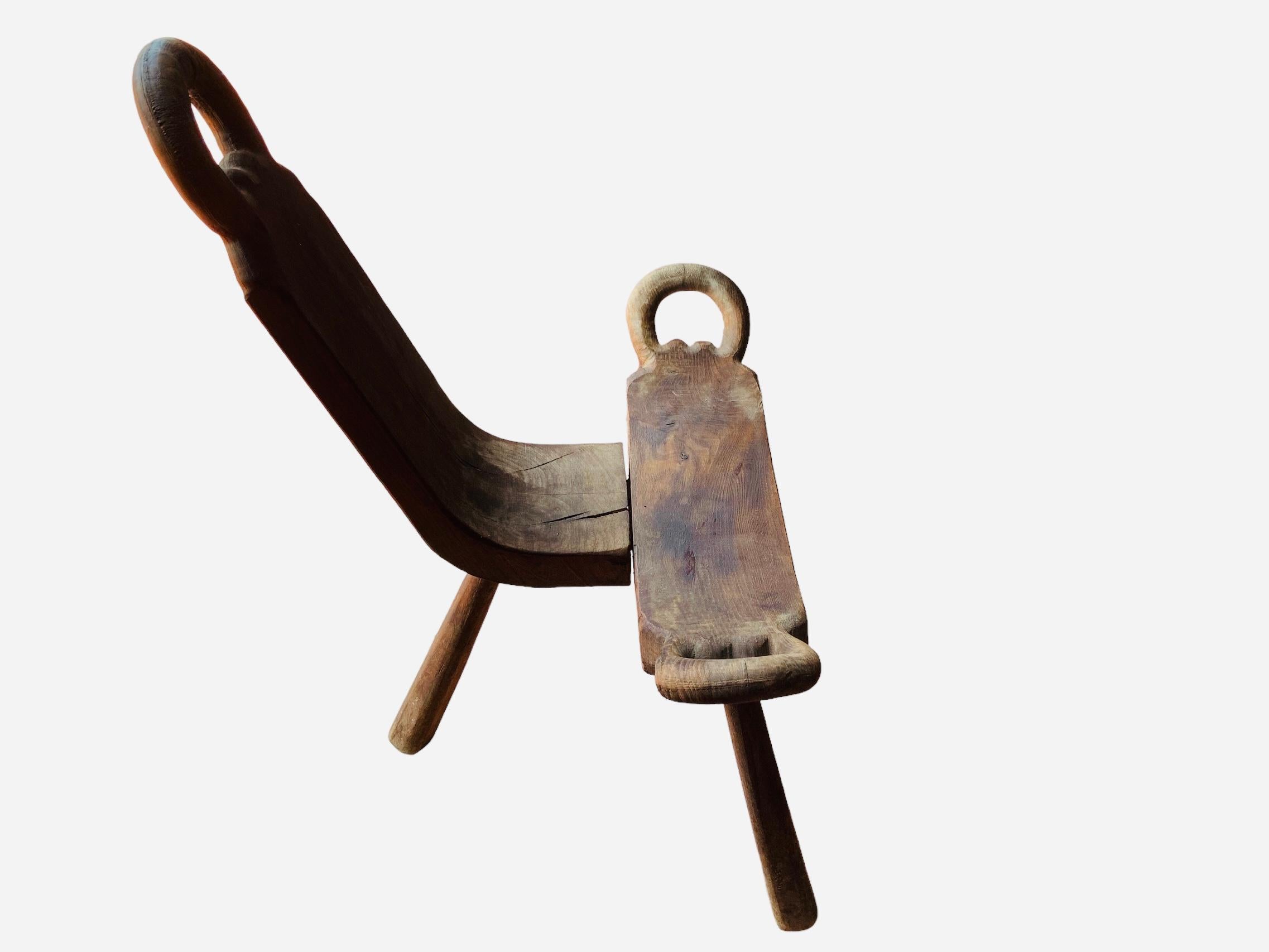This is a Spaniard Midwife Birthing Tripod Chair/Stool. It is made of solid wood. Its back is concave shaped and adorned at the top with a carved ring. The seat is rectangular like and also adorned with a ring at each side. The chair is supported by
